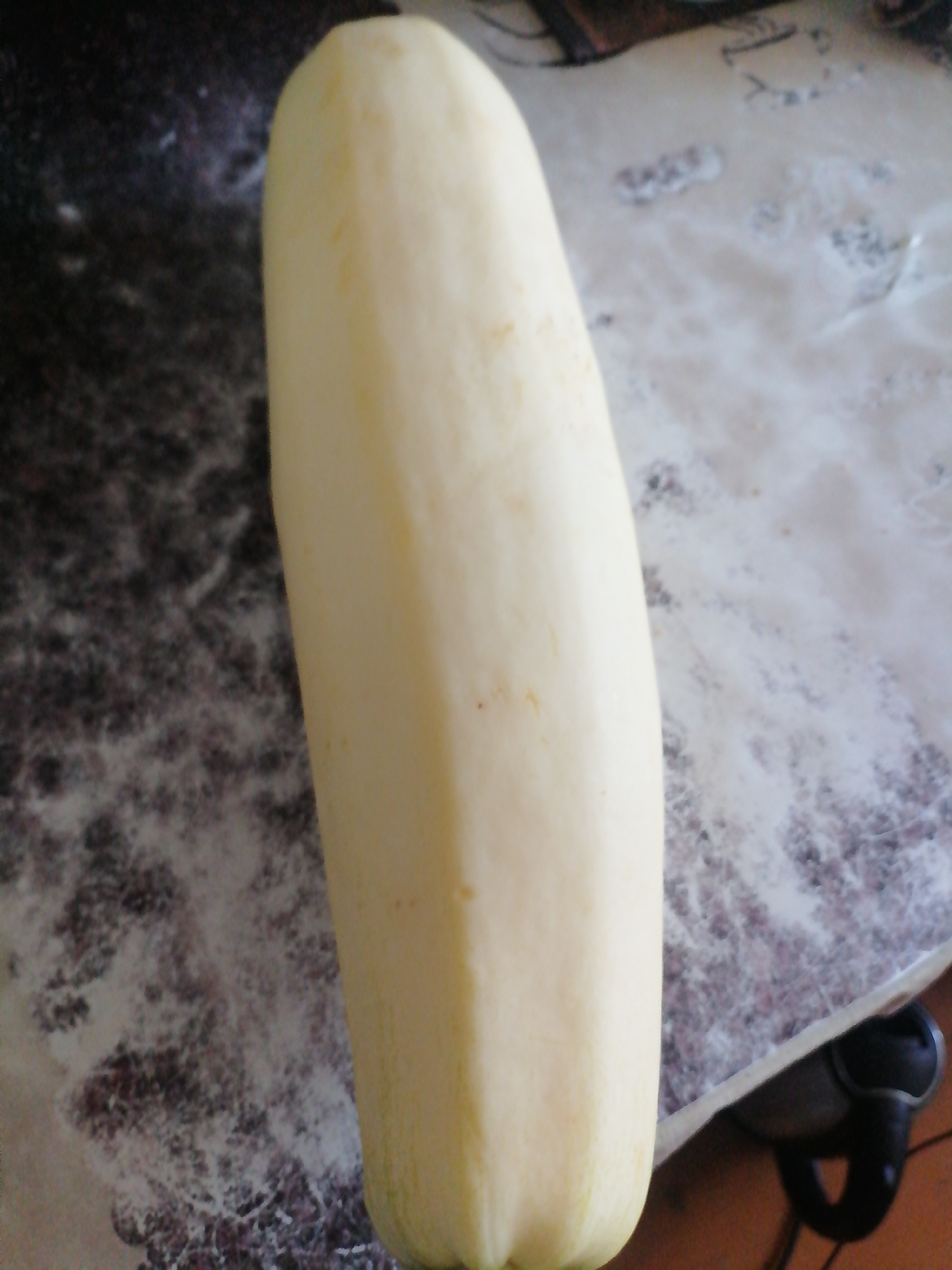 A pale yellow, oblong squash is centered on a patterned surface that appears to be a table or countertop. The squash is smooth and slightly curved, with subtle ridges running vertically along its length. The surface beneath the squash is dark with a speckled pattern, possibly granite or laminate. To the right of the squash, the surface transitions to a lighter, translucent material, possibly plastic wrap or a placemat, which also has a speckled pattern. In the lower right corner of the image, a dark object with a curved handle is visible, possibly part of a kitchen appliance or utensil. The lighting suggests an indoor setting.