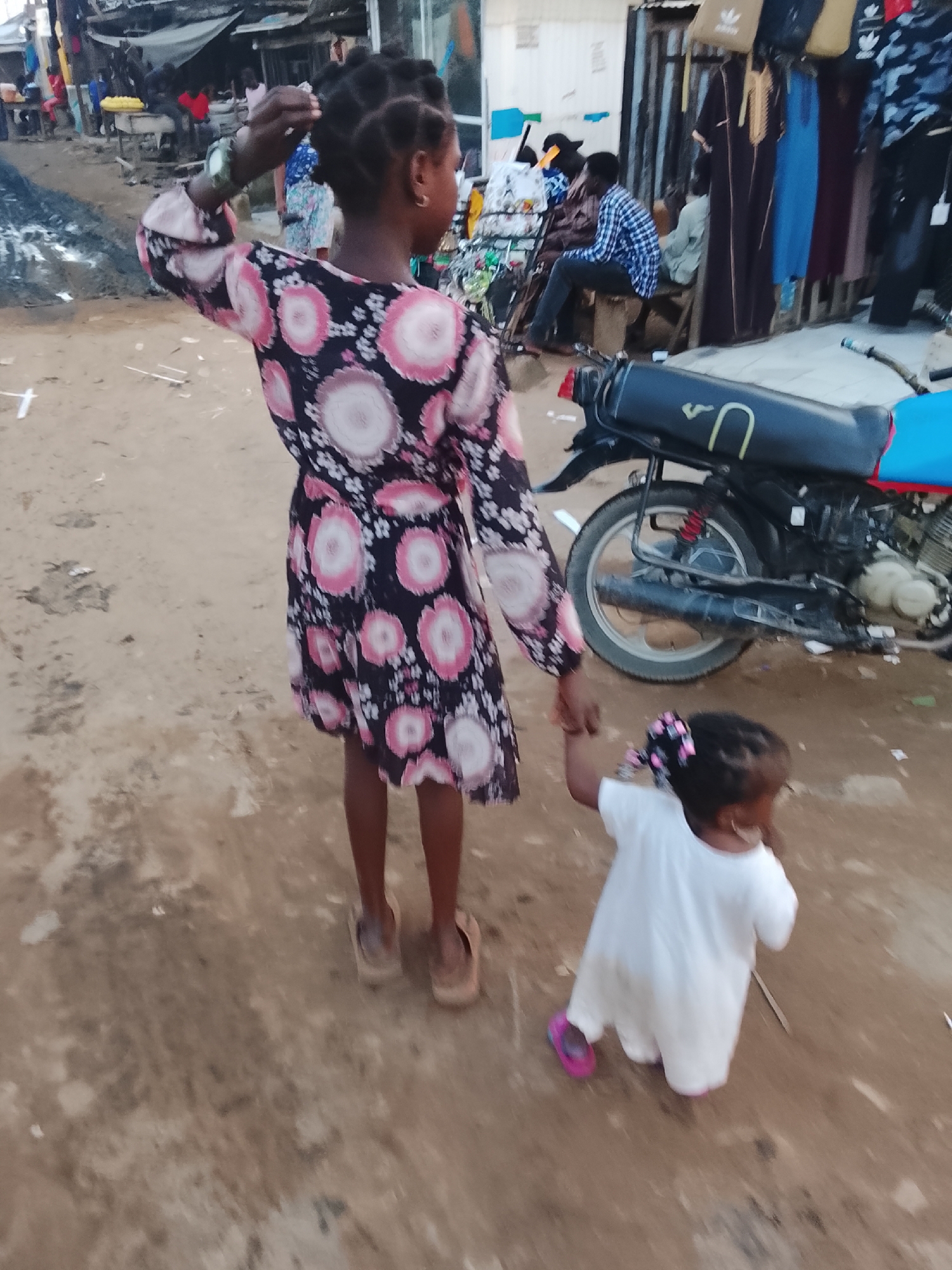 The image depicts two young girls in an outdoor marketplace environment, possibly in Abuja, Nigeria. A taller girl, facing away from the camera, wears a dark dress with a pink floral pattern and appears to be adjusting her hair. She has a watch on her left wrist. A younger girl, also facing away, is being held by the hand by the older girl. The younger girl wears a white dress and pink shoes, with a bow in her hair. They are standing on a dirt ground. In the background, there are market stalls with clothing hanging, a motorcycle, and several people, some of whom are seated. The overall scene suggests a busy, informal commercial area.