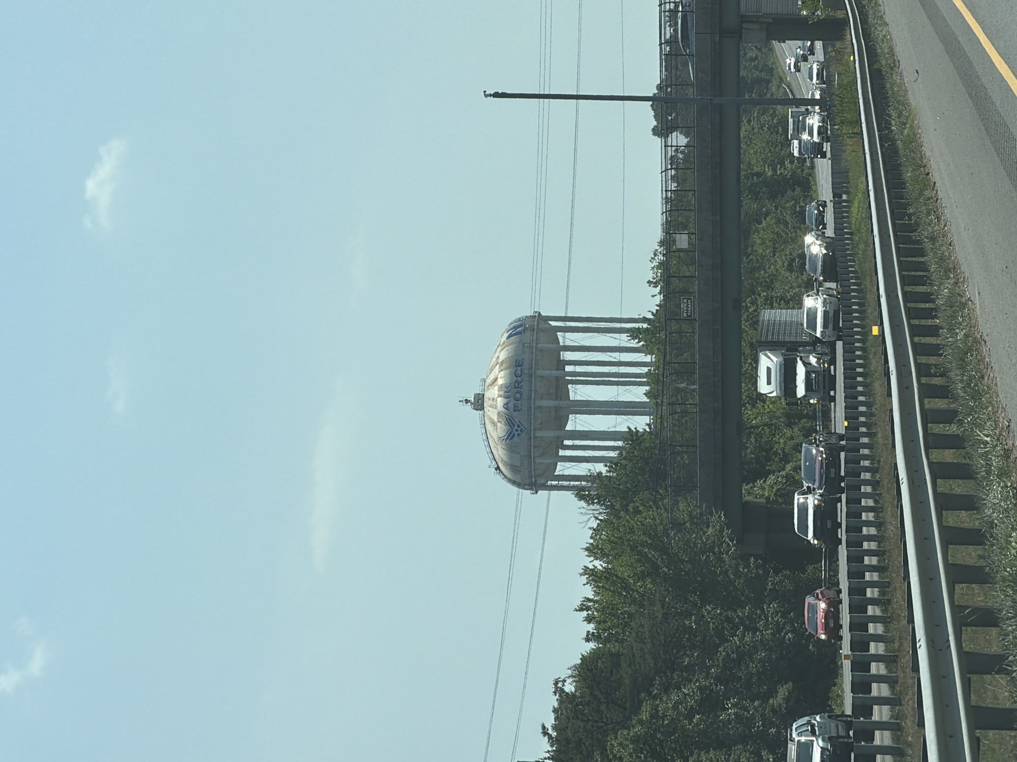The image shows a water tower with the United States Air Force logo and the word "FORCE" visible on its side. The water tower is a prominent structure in the background, set against a clear blue sky with a few wispy clouds. It is supported by a series of tall, thin legs. In the middle ground, a highway overpass with traffic is visible, with trees and vegetation lining the sides of the road. Several cars and trucks are driving on the highway. The foreground features a guardrail and some dry grass. The scene appears to be a roadside view in Forestville, United States of America.