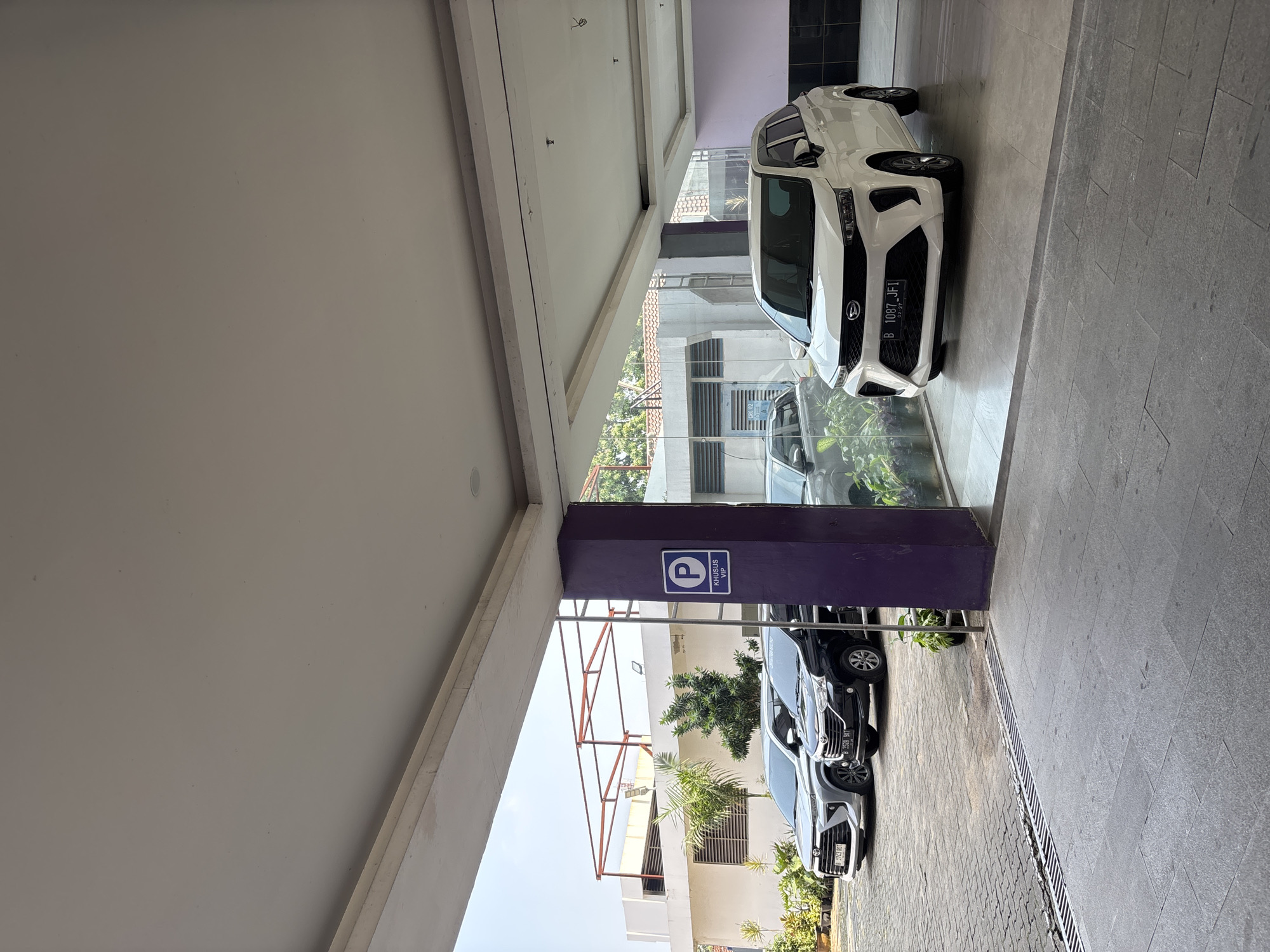 A white Daihatsu Xenia is parked in a covered outdoor area, likely a parking garage or building entrance, in Binong, Indonesia. The car has a license plate that reads "B 1087 JFI". The area is paved with large gray tiles and features a purple pillar with a blue parking sign indicating "KHUSUS VIP" (VIP ONLY). To the left of the white car, a black Daihatsu is parked, and further to the left, a silver SUV is visible. The background shows a building with windows and some greenery. The lighting suggests it is daytime with some sunlight.