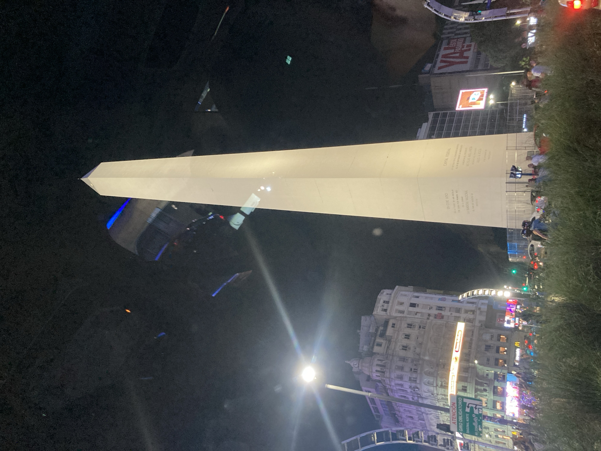 This nighttime shot captures the Obelisco de Buenos Aires in Argentina. The monument is illuminated, displaying inscribed text. People are gathered near the base of the obelisk, amidst greenery. The cityscape features buildings with lighted signs, and streetlights illuminate the area.