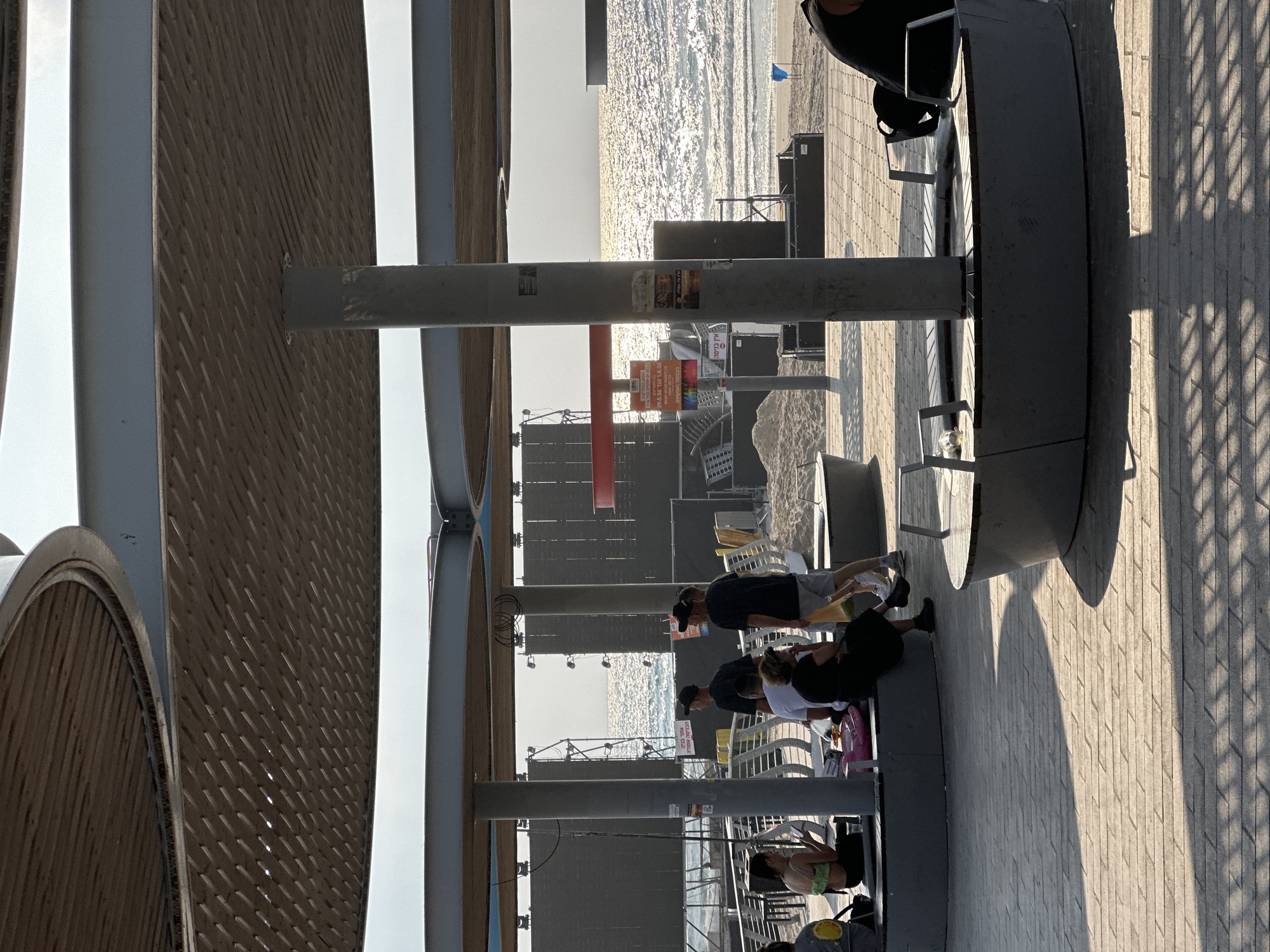 The image shows an outdoor paved area with several individuals seated on curved benches under a large, shaded structure. The structure has a wooden ceiling with a decorative, perforated pattern. In the background, the sea is visible with sunlight glinting on the water. There are also large, dark screens or stage backdrops erected on the beach.
Specifically, the location is on the Bat Yam promenade in Israel, a popular beachfront area.
On the left side of the image, a young woman with dark hair is sitting on a bench, wearing a green top and black pants, and holding a phone. Next to her, a person in a yellow shirt is also seated.
In the center, three men are standing under the structure. One man in a black t-shirt and black cap is facing away from the camera. Another man, also in a black t-shirt and black cap, is standing next to him, facing towards the sea. A third man is partially visible, wearing a black t-shirt and white shorts.
To the right, a woman is seated on a bench, wearing a black dress and black shoes. Another person in a black shirt is partially visible on the far right, seated.
Several benches are arranged around cylindrical support columns. The ground is paved with interlocking stones that create a patterned surface. Shadows from the structure are cast across the pavement, indicating the sun's position. In the distance, sand dunes and beach infrastructure are visible, suggesting the immediate proximity to the beach. Signs with Hebrew text are posted on the columns and near the beach. The overall atmosphere appears relaxed, with people enjoying the outdoor space near the sea.