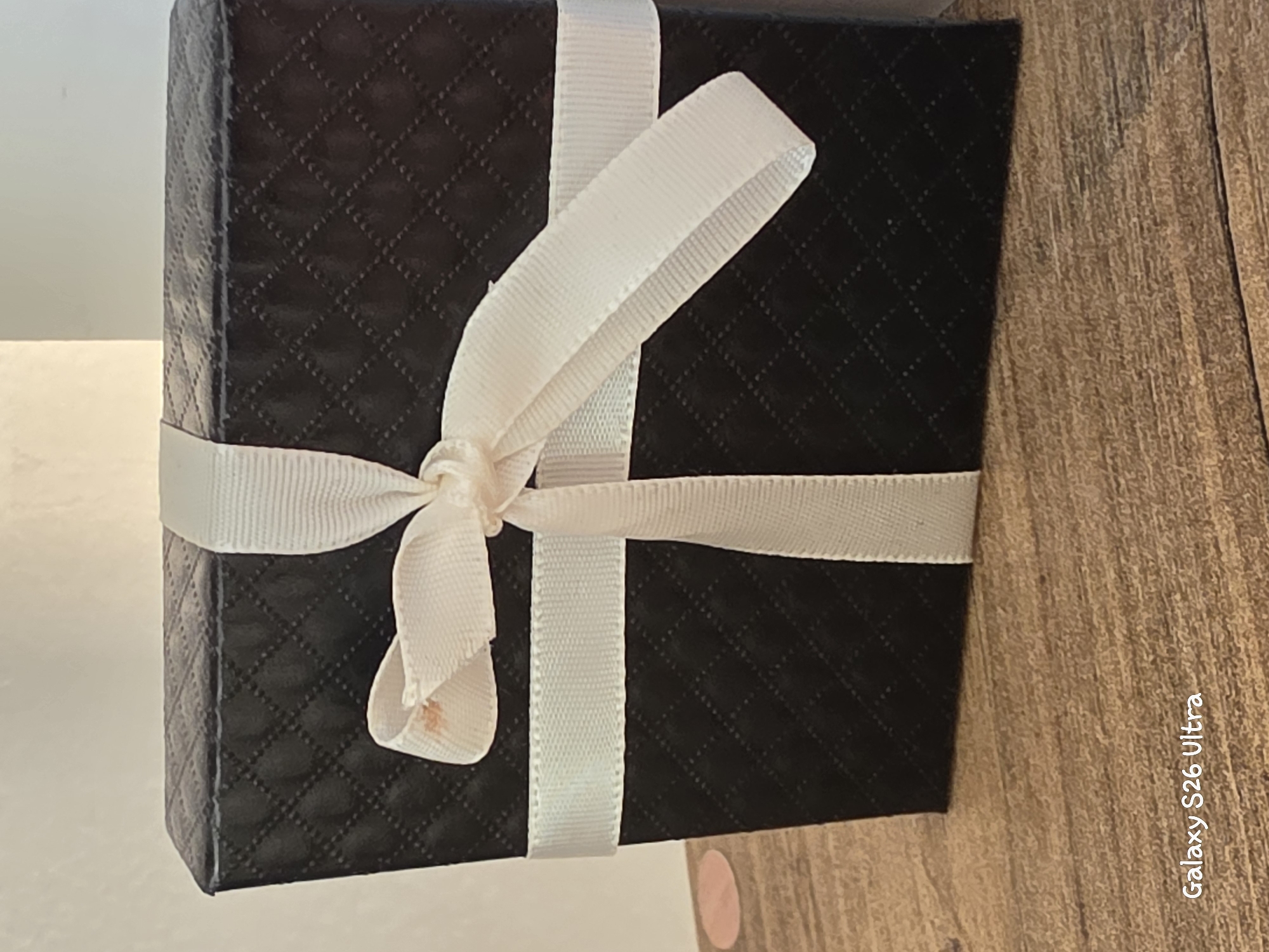 A black, quilted gift box tied with a white ribbon sits on a wooden surface. The ribbon is tied in a bow on the front of the box, with one loop of the bow showing a faint reddish-brown smudge. The surface beneath the box is a textured wood grain. In the lower left corner of the image, text reads "Galaxy S26 Ultra". The background is a plain, off-white wall. There are no people or animals present. The image does not provide any information about a location in Plovdiv, Bulgaria.