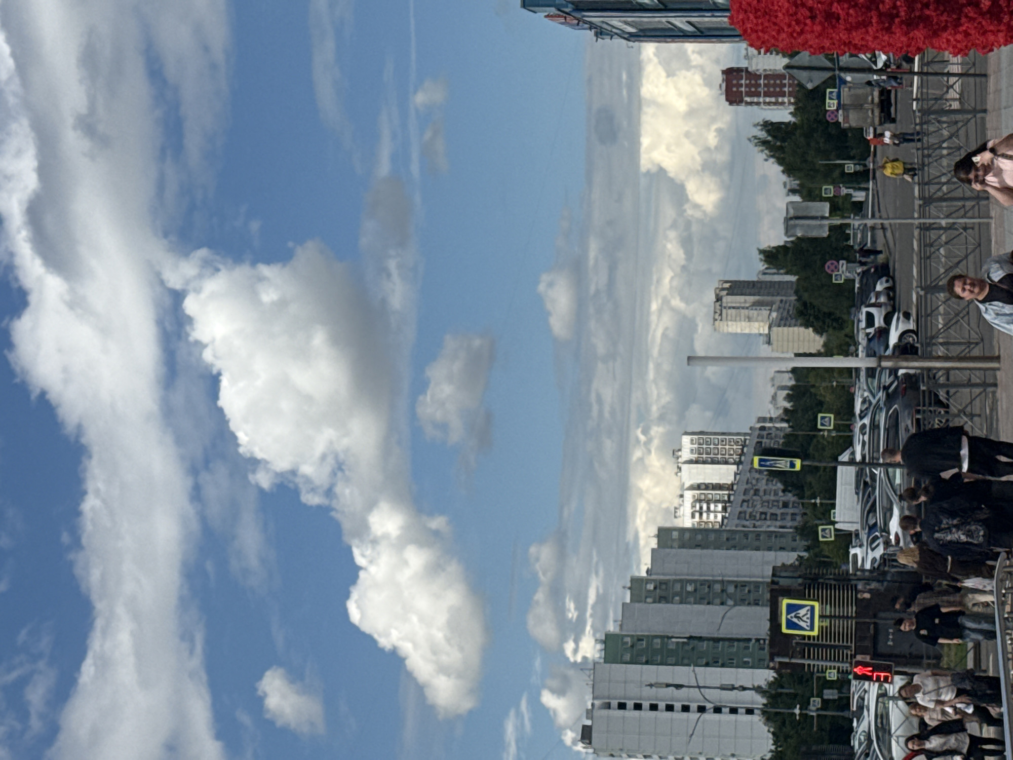 This is a full shot of a street scene in Moscow, Russia. The image is taken from a low angle, looking upwards towards a bright blue sky with scattered white clouds. Buildings of various heights line the street, some appearing to be residential high-rises with many windows. The street itself is lined with parked cars and a sidewalk where several people are walking. Traffic lights and pedestrian crossing signs are visible. A large, textured red object is visible at the top left of the frame, possibly a decorative element or artwork. The overall impression is a typical urban environment on a clear day. The time of day appears to be daytime, likely late morning or afternoon, given the brightness of the sky. The weather is fair. The emotions of the people are not clearly discernible due to the distance and resolution. No visible text is legible in the image.