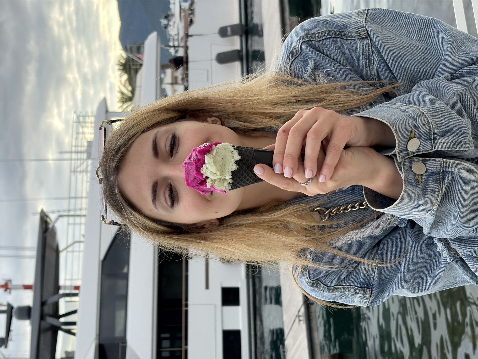 A blonde woman wearing a denim jacket holds an ice cream cone with two scoops, one pink and one light green, in front of her face. She is standing in Tivat, Montenegro, based on the background elements. The background features a marina with several yachts and boats, a partly cloudy sky, and distant mountains. The woman is wearing glasses propped up on her head. She has a ring on her finger. The water from the marina can be seen in the foreground.