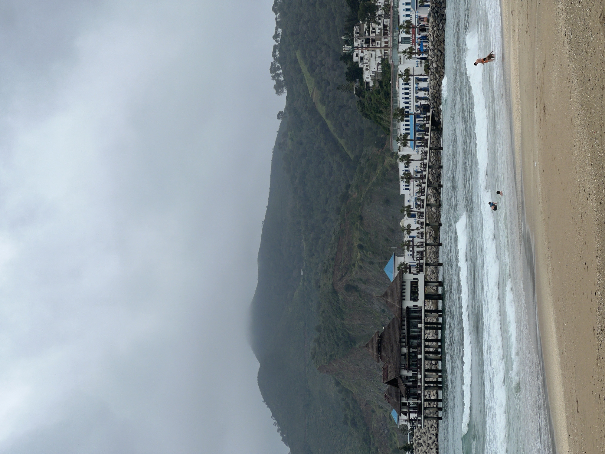 This image captures a picturesque coastal landscape in M'Diq, Morocco, characterized by a sandy beach, ocean, and mountainous backdrop under an overcast sky.
In the foreground, a wide, light-brown sandy beach occupies the right side of the frame, meeting the turquoise-grey waters of the ocean. Several gentle waves are breaking and rolling towards the shore. A few individuals are visible in the water; one person wades in the shallow surf, while another is further out where the waves are more prominent.
The midground features a dense row of white and blue multi-story buildings, likely hotels or resorts, lining the coast. Palm trees are interspersed among these structures. A prominent pier-like structure with dark brown roofs and multiple pillars extends into the water from the shoreline, suggesting a restaurant or recreational facility. A rocky breakwater, possibly constructed with tetrapods, is also visible along parts of the coastline, separating the buildings from the sea.
Dominating the left side and background is a large, verdant mountain range, thickly covered with green vegetation and trees. The upper reaches of the mountain are partially shrouded in clouds or mist, contributing to the soft, diffused light and an atmospheric sense of depth. The sky itself is entirely overcast, suggesting a cloudy or foggy day.
The scene depicts a tranquil coastal setting, with people enjoying the ocean, set against a dramatic natural and developed backdrop. There is no visible text in the image.