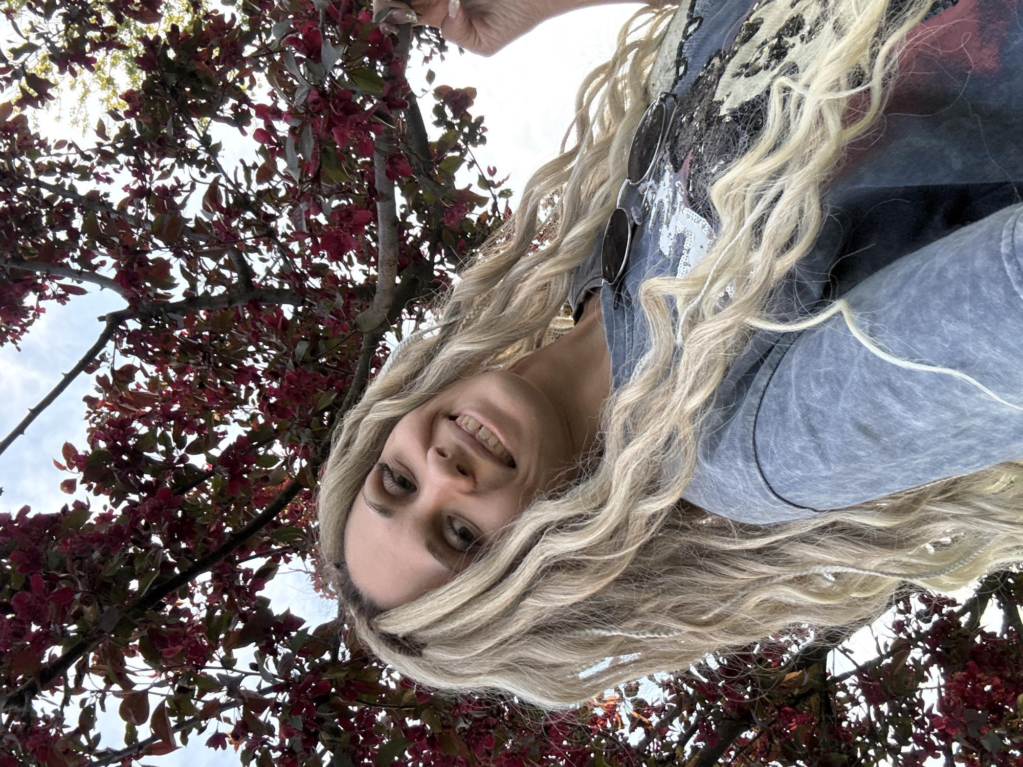 A woman is pictured from a low angle against the backdrop of a tree with red leaves and a cloudy sky. She has long, blonde, wavy hair with a few braids and is smiling. She is wearing a gray top, with sunglasses hanging from the collar. The photograph was taken outdoors. The location details have not been provided in this image.