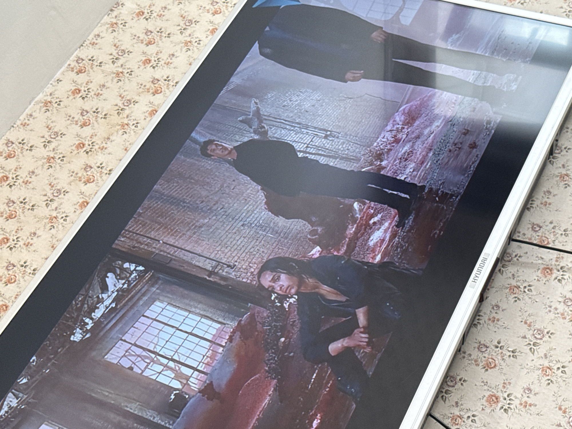 The image shows a scene displayed on a Hyundai monitor. The scene appears to be from a movie or TV show set in a dilapidated, possibly post-apocalyptic environment. There are two main characters visible: a woman crouching down and a man standing. Both are wearing dark clothing. There is significant damage and what appears to be blood splattered around the room. The environment looks like an abandoned building with broken windows and decaying walls. The location details are not visible in the image.