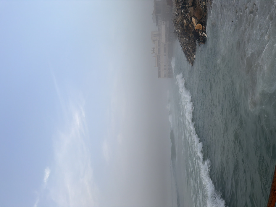 A landscape-oriented image showcases a hazy, overcast scene. The left side is dominated by a pale blue sky, blending into thick fog. To the right, choppy water creates foamy waves near a bank cluttered with debris, possibly rubble and vegetation. In the distance, a building emerges faintly through the mist. The overall atmosphere is muted and obscured by fog.