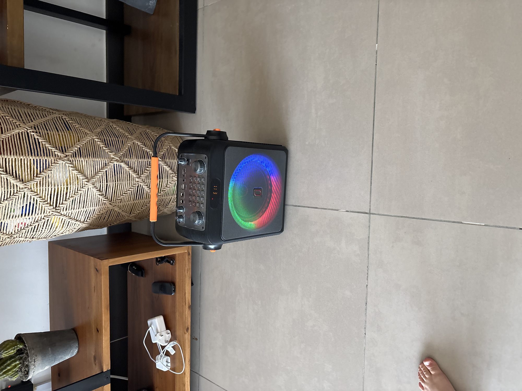 The image captures a portable speaker with a colorful LED light display, positioned on a tiled floor. The speaker is situated in the foreground, to the right of a wooden shelf unit. On the shelf, various items are visible, including electrical cords, a power adapter, and a small black device, possibly a remote control or portable hard drive. To the left of the speaker, a large woven basket stands next to the shelf. In the upper left corner, a potted plant with green leaves is visible, resting on top of the shelf. A person's foot, with painted toenails, is visible in the bottom left corner of the image. The scene appears to be indoors, possibly in a residential setting, with neutral-colored tiled flooring and a plain background wall. The lighting suggests an indoor environment, possibly with natural light contributing to the overall illumination. The image does not provide any specific landmarks or contextual clues to indicate its location within Dubai, United Arab Emirates.