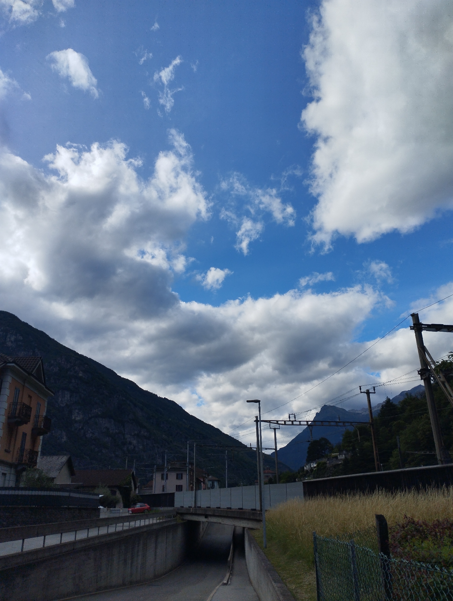 An elevated, eye-level, full shot captures a cloudy, bright day in a mountainous area with some structures and a road. The sky is a bright blue with scattered, fluffy white and gray clouds. In the foreground, a concrete structure leads into a tunnel. A chain-link fence runs along the right side of the image, partially obscured by dry grass and some bushes. A single red car is visible in the distance, driving on a road. To the left, a building with orange and white walls and balconies sits at the base of a dark, rocky mountain. In the background, more buildings and trees are visible, with a train track and power lines running across the scene. The overall scene is serene and industrial, with the natural beauty of the mountains contrasting with the man-made structures.