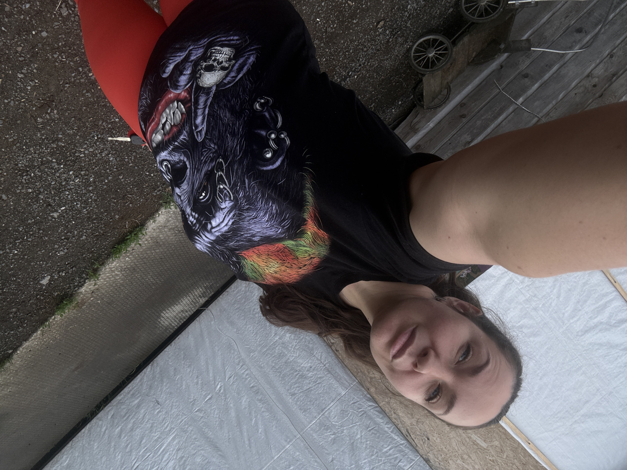 A woman is lying down on her back with her head slightly tilted to her left. She is wearing a black t-shirt with a graphic print of a wolf's head and a skull, and red leggings. Her eyes are open and she is looking directly at the camera. The background is a mixture of dirt, gravel, and a wooden fence. There are also two old wheels visible in the background. The image appears to have been taken outdoors. The location in Chaikovsky, Russia, is not discernable from the provided image.