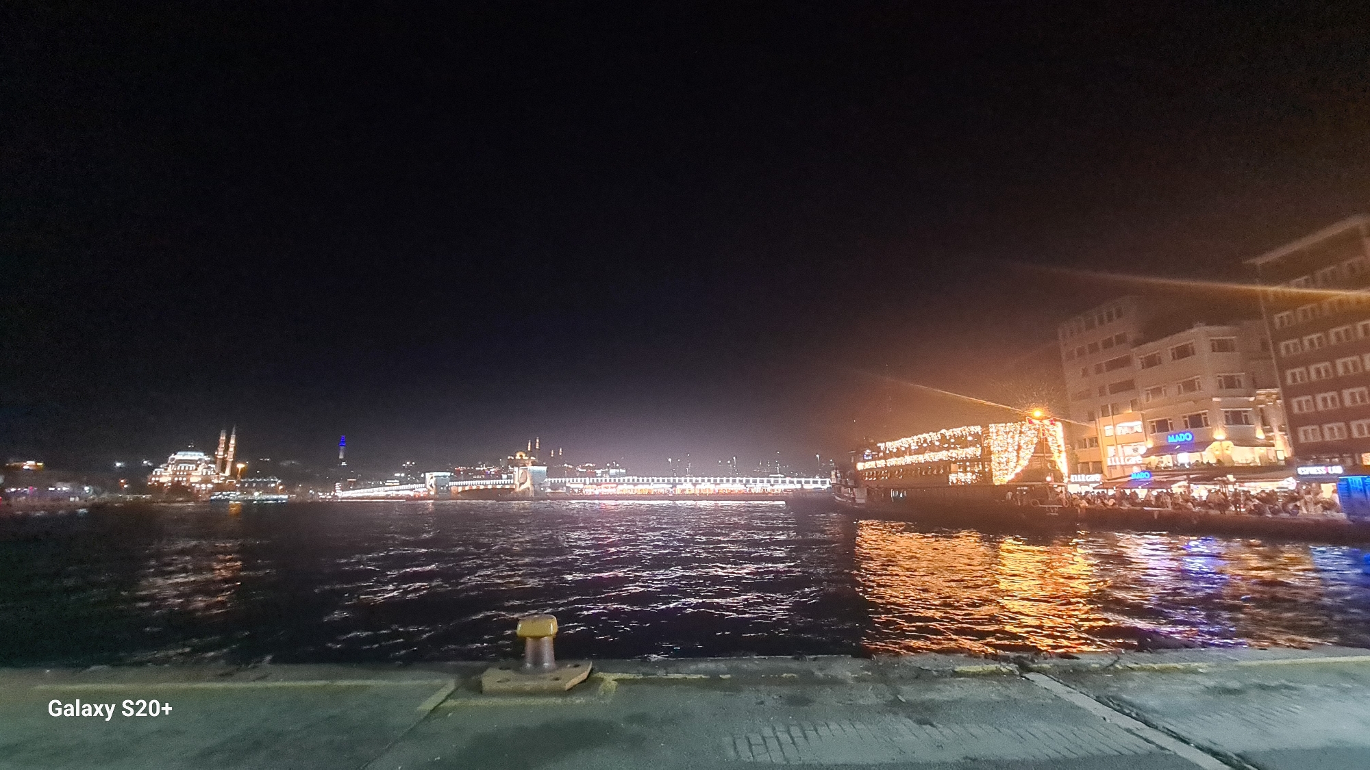 This is a night-time, wide-angle photograph taken from the shore, likely in the Beyoğlu district of Istanbul, Turkey, looking across a body of water. The scene is illuminated by numerous artificial lights.
On the left side of the frame, a minaret and a domed structure, possibly a mosque, are visible in the distance, lit up against the dark sky. Further along the waterfront, a long bridge, likely the Galata Bridge, is visible, its lower sections lined with lights.
Dominating the right side of the frame is a large boat, heavily decorated with strings of bright lights, creating a festive appearance. People are gathered on the deck and surrounding areas of the boat. Behind the boat, several buildings with illuminated windows are visible, including one with a prominent "MADO" sign.
The water in the foreground reflects the lights from the boat and the buildings, creating shimmering patterns. In the lower left corner, the text "Galaxy S20+" is present, indicating the device used to capture the image. A yellow bollard is visible in the immediate foreground. The overall atmosphere is vibrant and lively, suggesting a bustling night scene.