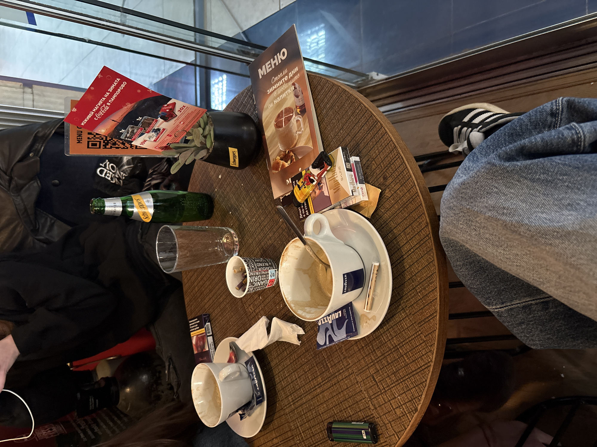 The image captures a cluttered coffee table scene, likely in a café or restaurant in Pleven, Bulgaria. The table is round with a dark brown woven pattern. Two people are partially visible, one with a black jacket and the other with blue jeans and Adidas sneakers.
On the table are two used coffee cups with saucers, an empty Schweppes bottle, a drinking glass, and a small paper cup. There are several packs of cigarettes (Marlboro, Ome) and a lighter. A menu card stands upright, next to a Coca-Cola promotional card.
The environment appears to be an indoor café setting with windows and a metal railing in the background. The scene suggests a casual, everyday moment of coffee and conversation.
