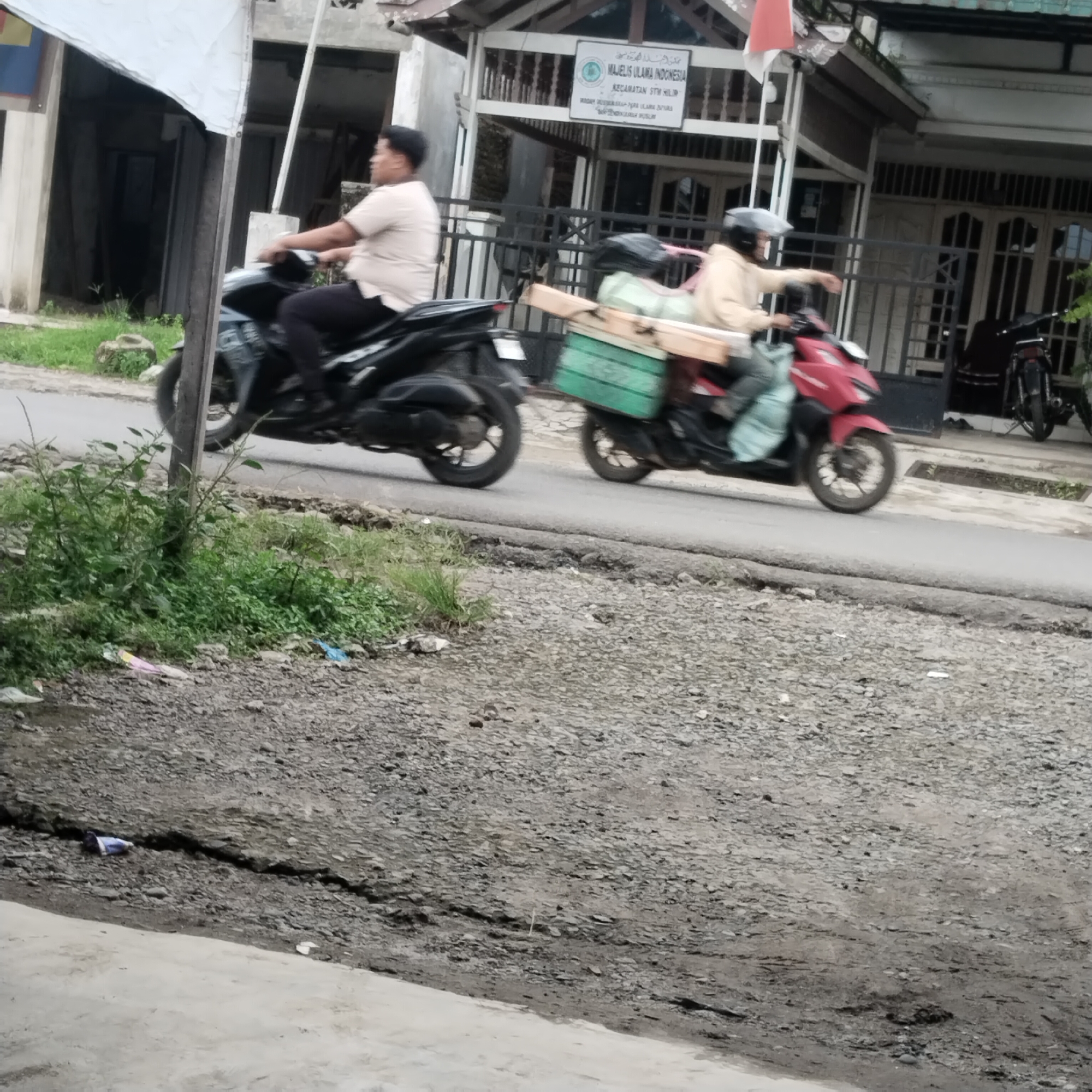 Two motorcyclists are traveling on a paved road in front of a building with a sign that reads "MAJELIS ULAMA INDONESIA KECAMATAN SUMPIUH". The motorcyclist on the left is a person wearing a light-colored t-shirt and dark pants. They are riding a black motorcycle. The motorcyclist on the right is wearing a light-colored jacket and a helmet, and is also wearing a helmet. This motorcycle is red and has green and brown cargo boxes attached to the rear. The ground is covered with gravel and patches of grass. There is a crack in the paved road in the foreground. The scene appears to be in a semi-urban or rural area of Talun Kenas, Indonesia, given the building and the road conditions.