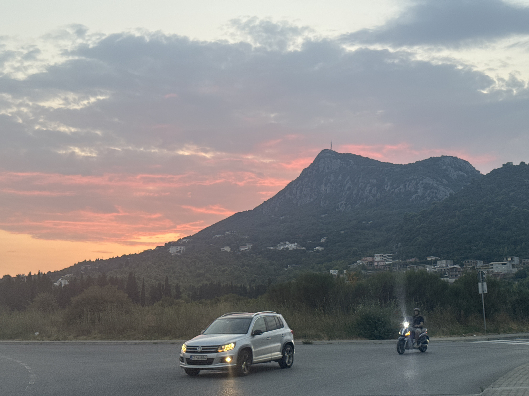 In Sutomore, Montenegro, as the sky transitions to dusk with soft pink and orange hues, a silver Volkswagen Tiguan is parked on an asphalt road. The SUV's headlights are illuminated. To the right of the Tiguan, a person is riding a dark-colored scooter with its headlight on, also on the asphalt. In the background, a lush green mountainside is dotted with buildings, and a few sparse trees are visible. The sky is filled with a mix of light and dark clouds.