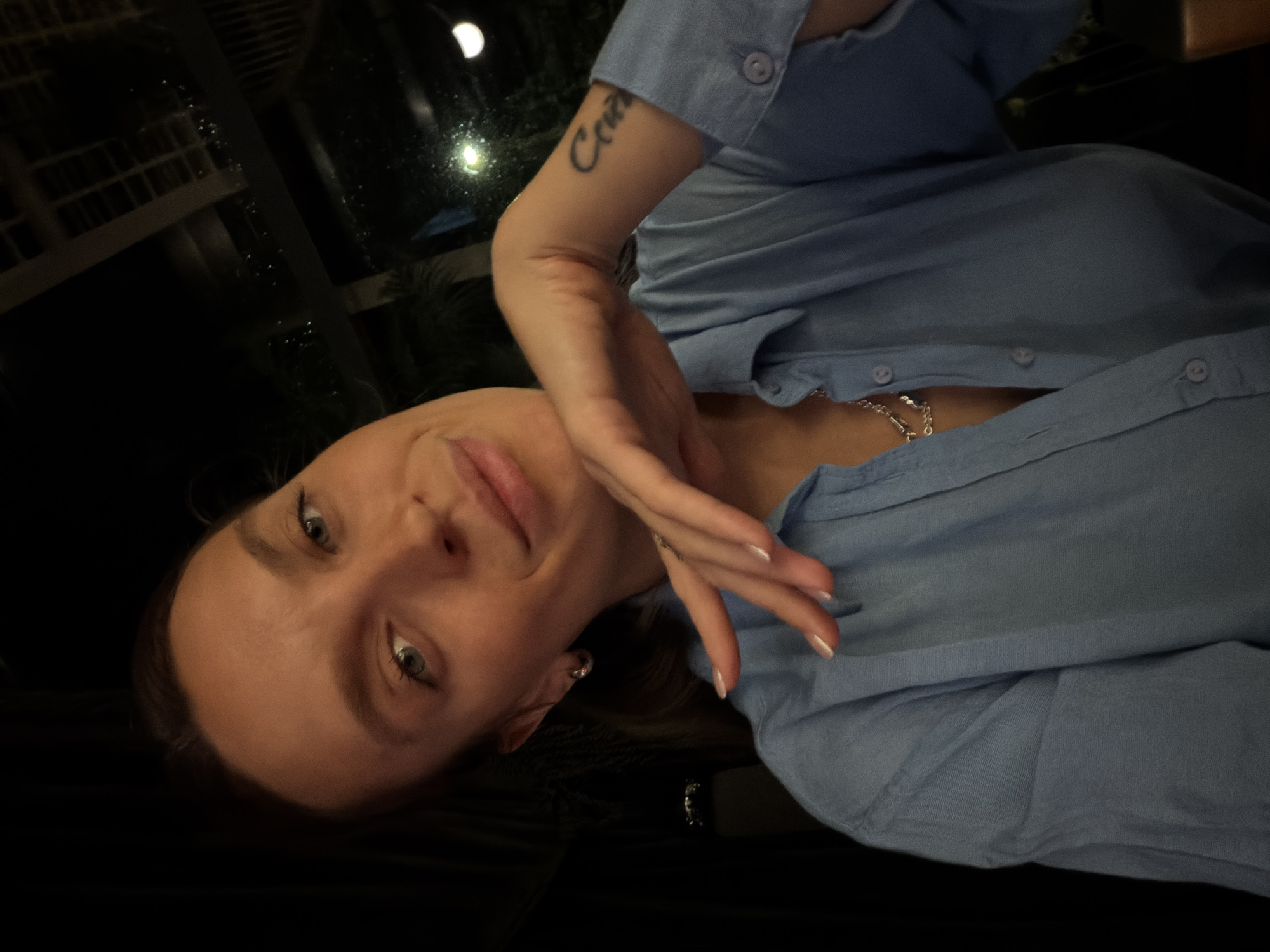 The image is a portrait of a young woman, captured from a rotated perspective, giving the impression she is lying down or leaning back. She is looking directly at the camera with a soft, engaging smile. Her light-colored eyes are prominent, and her dark hair is visible against a dark background.
She is wearing a light blue, collared, button-down shirt. The shirt is partially unbuttoned, revealing a silver chain worn around her neck. Her left hand is positioned gracefully near her face, showcasing a neat French manicure with white-tipped nails. On her right forearm, a tattoo in stylized script spells the word "Cool." A small silver hoop earring can be seen in her left ear.
The background is predominantly dark, indicating a nighttime setting or a dimly lit indoor space. Reflections are visible on what appears to be a window or glass, showing faint lights from outside, including a crescent-shaped light and other distant artificial illumination, suggesting an urban or built-up environment at night. The overall mood is relaxed and casual. The location context for this scene is Budva, Montenegro.