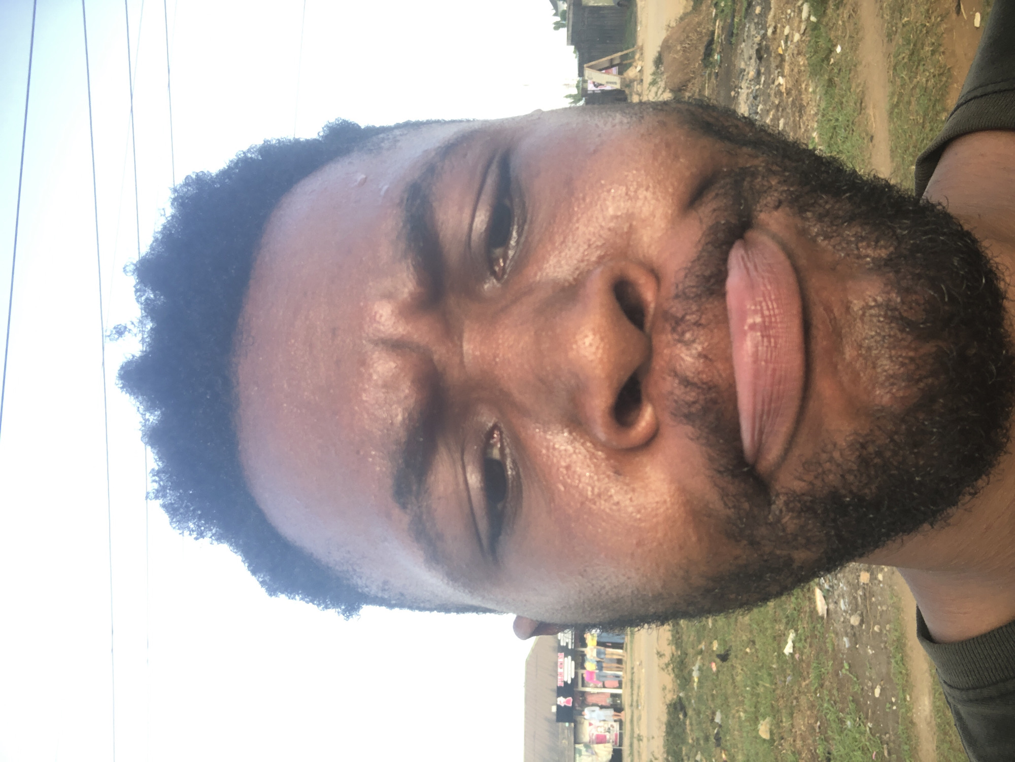 A close-up, vertically oriented photo shows a Black man with a full beard and close-cropped hair. His face appears slightly sweaty. He has his tongue sticking out. In the background, there is a glimpse of a street scene with buildings and greenery. Utility lines are visible in the upper left of the frame. The location is Unknown, Unknown.