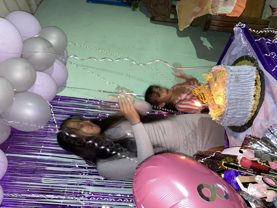Two people lie on a shiny, purple surface among birthday decorations. To the left are silver and purple balloons. A woman with long, dark hair wears a light grey dress. A child in a striped shirt lies next to a birthday cake decorated with lit candles. A large pink balloon with a silver number "8" is in the foreground. The background shows a green wall and a wooden chair. Gifts and purple and white decorations fill the scene.