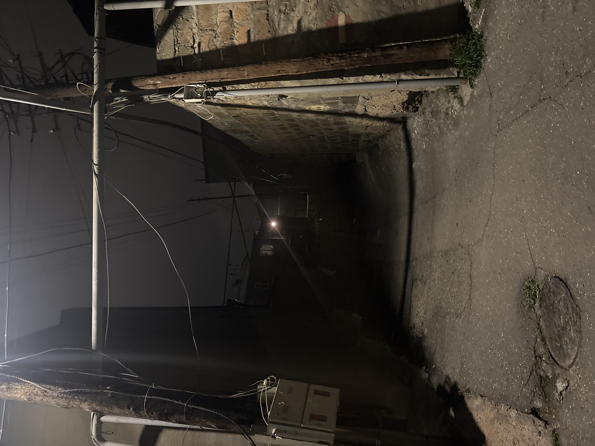 This is a night shot of an alleyway somewhere in Yerevan, Armenia. The alley is narrow, with brick or concrete walls on either side. It is paved with asphalt, which is cracked and worn in places. There's a manhole cover on the right side of the alley floor. There is exposed wiring and a utility pole on the left side of the frame. A light source is visible down the alley, suggesting the presence of street lights or buildings at the end of the alleyway. The light reflects on the pavement. There is a bit of vegetation near the walls of the alley. The scene is dimly lit, giving it a somewhat dark and gritty feel.
