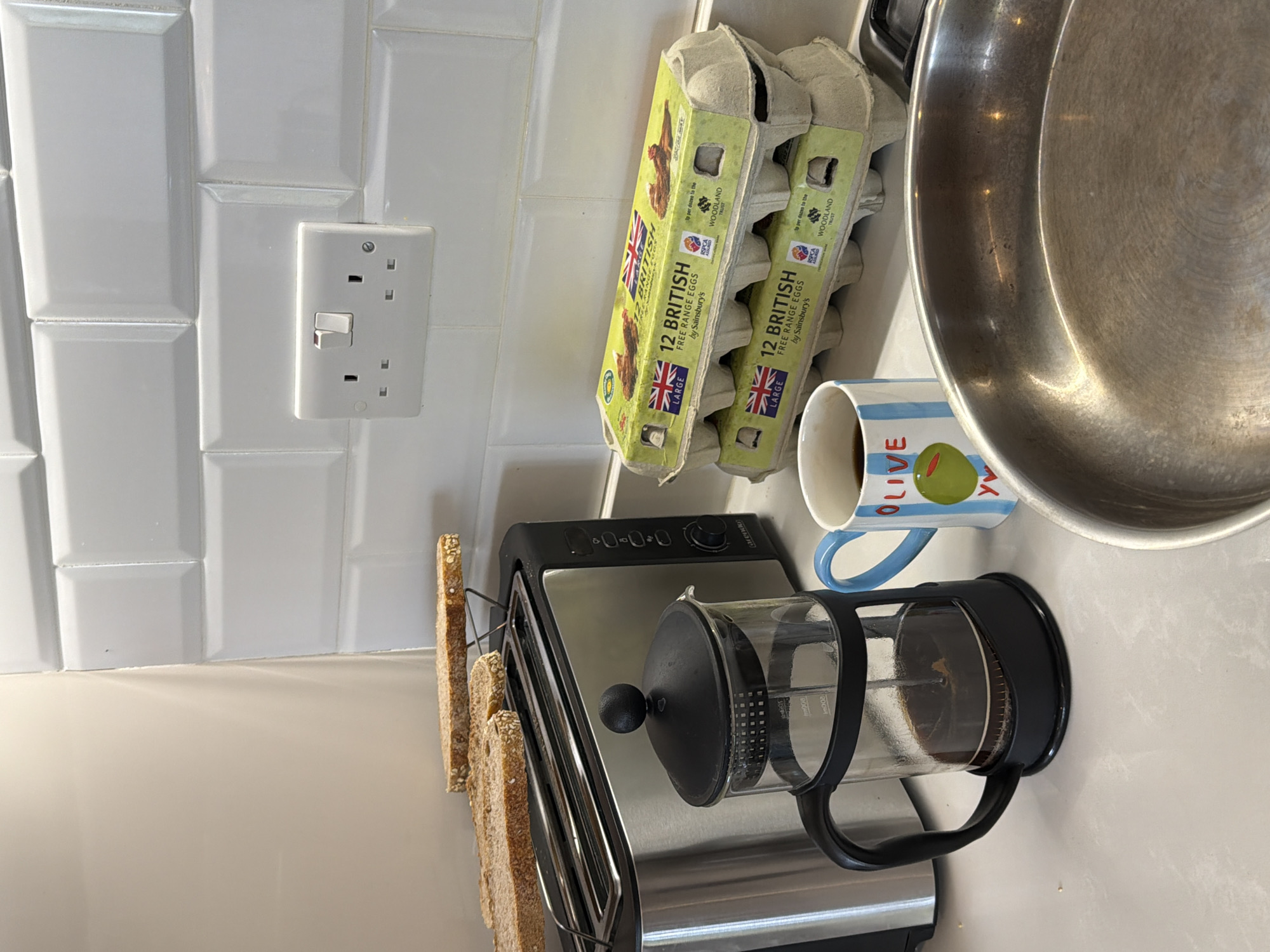Here's a detailed description of the image:
**Objects:**
* Two cartons of "12 British Free Range Eggs by Sainsbury's" are visible, indicating a UK origin.
* A mug with the word "OLIVE" and a picture of an olive is present.
* A French press filled with coffee.
* A silver frying pan.
* A toaster containing toast.
* A white double electrical outlet.
**Environment:**
* The background features white, tiled walls, typical of a kitchen or food preparation area.
* The countertop is white.
**Inferred Location:**
* The image is likely from a kitchen in the UK, based on the "British Free Range Eggs" packaging. However, there is no indication of the city of London, United Kingdom.