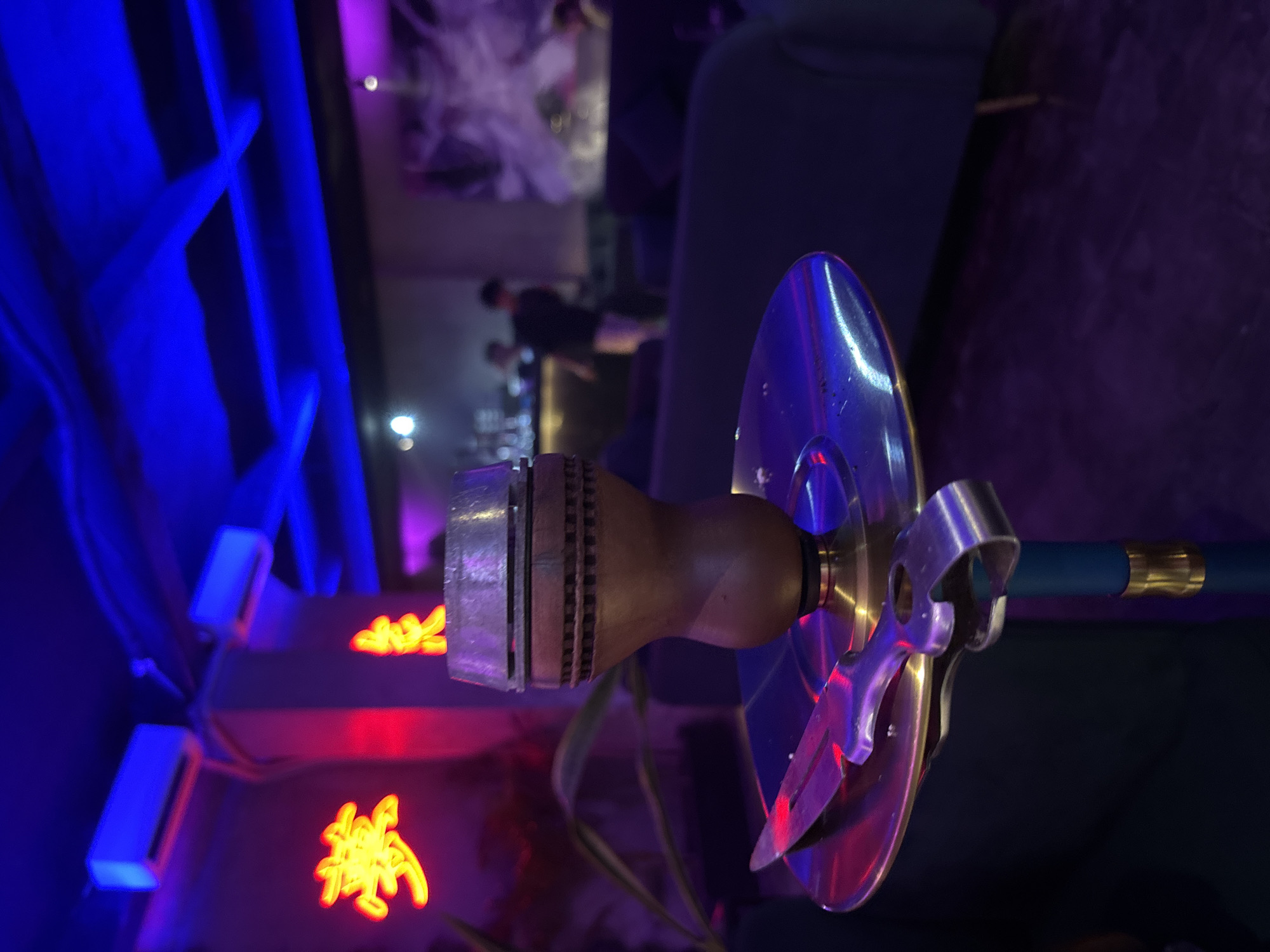 This image captures a vibrant and dimly lit indoor setting, likely a shisha lounge or bar in Kuta Selatan, Indonesia. The main subject, positioned prominently in the foreground, is the top section of a hookah (shisha) pipe. It features a wooden stem or bowl holder, a metallic ash tray, and a pair of silver tongs hooked onto the tray, suggesting it is either in use or ready to be used. A dark blue hose with a gold-colored fitting extends to the right.
The background is out of focus, enhancing the depth of field and drawing attention to the hookah. Blue neon lights cast a strong glow on the left side of the frame, illuminating structural elements like vertical slats. Further back, red neon light highlights what appears to be a stylized sign or text, though it is too blurry and intricate to be legible. Blurry figures of people are visible in the deeper background, suggesting a social gathering. Furniture, possibly dark chairs or sofas, can also be discerned in the soft-focused backdrop.
The artificial lighting, characterized by the strong blue and red neon glows and reflections on the metallic surfaces of the hookah, indicates a nighttime or evening setting. The overall atmosphere appears relaxed and social, typical of a lounge environment.