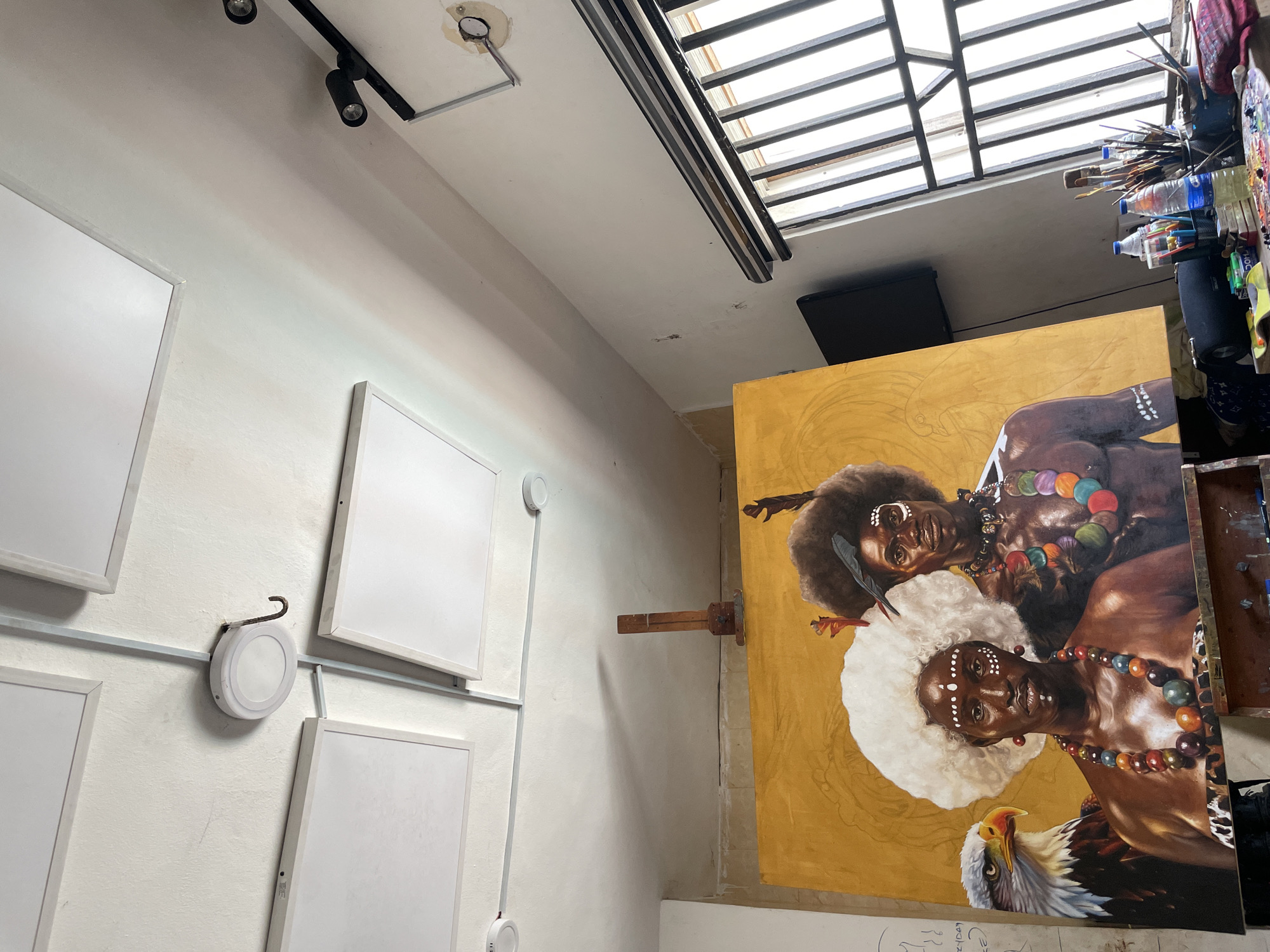 The image shows the interior of what appears to be an artist's studio. On the left wall are several framed white canvases arranged vertically. A white cable runs along the wall, connecting small, round, white devices. Above, there is a black track lighting fixture.
On the right side of the image, a large canvas stands upright, depicting two dark-skinned individuals adorned with colorful jewelry and headwear, along with an eagle at the bottom. The background of the painting is gold. Art supplies, including brushes and bottles, are visible to the right of the canvas.
Above the canvas, a skylight is present. A black object, possibly a speaker, is mounted nearby.
It is not possible to determine the specific location within the city of Ado, Nigeria based on the image alone.