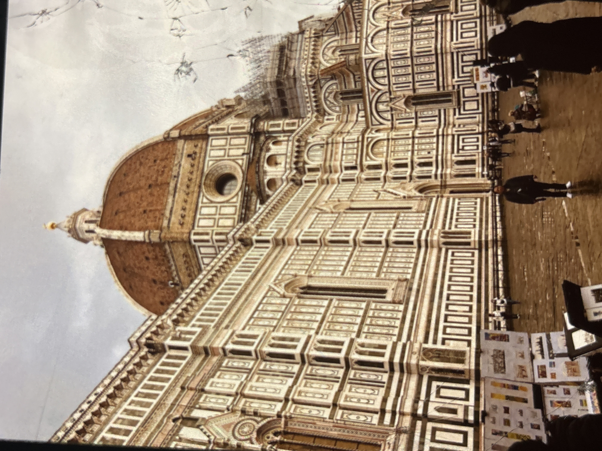 The image depicts the Florence Cathedral (Duomo di Firenze) in Florence, Italy. The architectural style is Renaissance, characterized by its prominent dome, intricate facade with marble inlay, and numerous arched windows. In the foreground, several people are visible, some appearing to be tourists or locals walking on the wet piazza in front of the cathedral. There are also stands set up displaying what appear to be artwork or souvenirs. The sky is overcast. The provided description about Orihuela, Spain is incorrect; the landmark is in Florence, Italy.