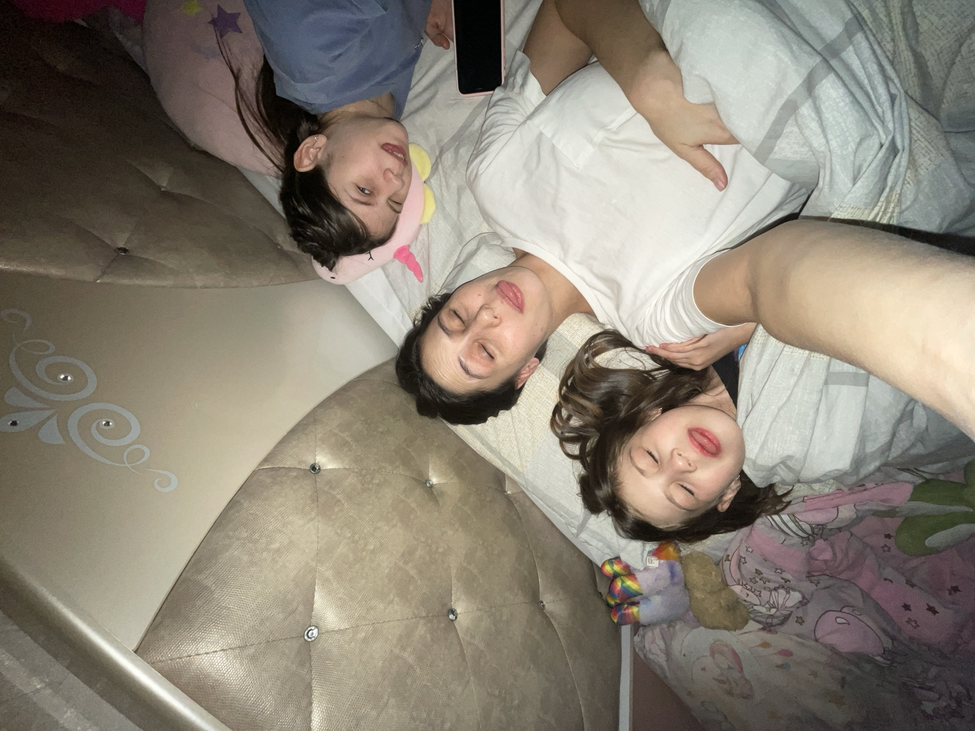 The image displays three females lying in a bed within a domestic indoor setting. The specific location in Izhevsk, Russia, cannot be determined from the visual evidence.
In the center foreground, a female with dark hair, wearing a white short-sleeved t-shirt, lies on her back. Her eyes are closed, and her tongue is partially extended. Her right arm, cropped at the elbow, extends towards the viewer, suggesting she is holding the camera.
To her left, a younger female child with dark hair, wearing a dark-colored shirt, lies with her head resting on the first female's left arm. Her eyes are closed, and her tongue is also partially extended, mirroring the first female. She is partially covered by a pink blanket featuring unicorn and star patterns. Several colorful plush toys, including a rainbow-colored item, are visible on the bed near her.
To the right and slightly above the central figure, a third female, appearing to be an older child or young adult with dark hair, lies on her back. She wears a blue long-sleeved shirt and rests her head on a pink unicorn-shaped pillow with a yellow flower detail. Her right eye is closed in a wink, and her mouth is slightly open. A dark-colored smartphone with a pink case lies face-down on the bed next to her head.
The bed features a large, light-gold or beige tufted headboard with a textured finish and silver-colored button accents. Above the headboard, a curved wall or architectural detail is visible, adorned with silver-colored swirl patterns and small embedded decorative elements. The bedding is light-colored, with a grey patterned duvet partially covering the central and left figures. The lighting suggests an indoor, possibly dim or evening, environment.