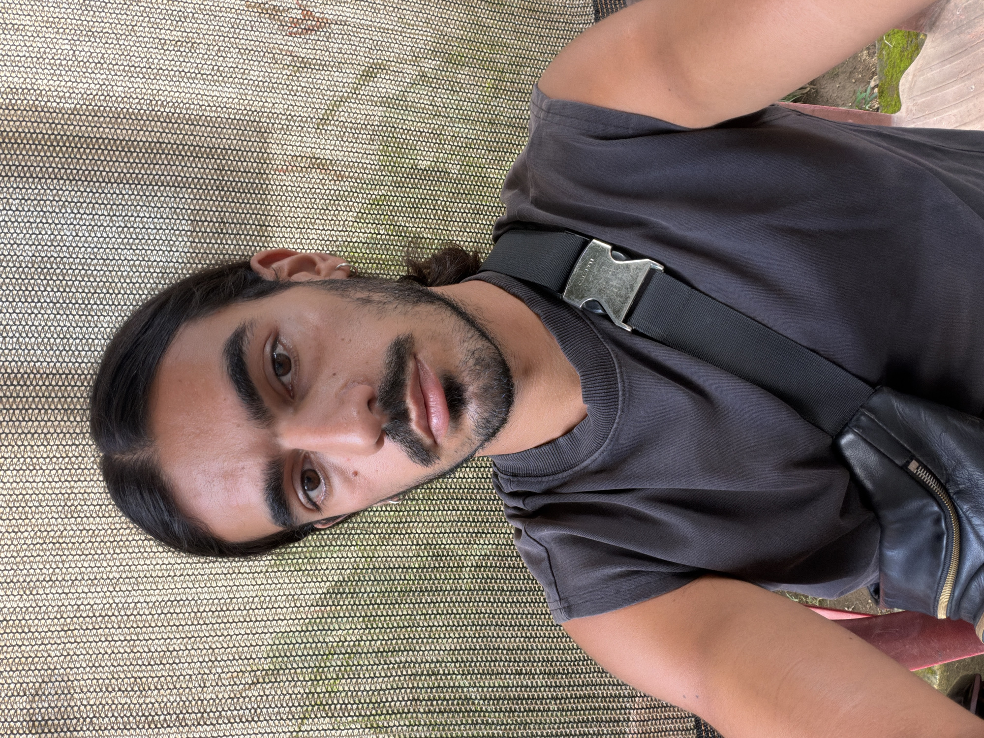A man with dark hair, a beard, and brown eyes is seated on a surface with a fine mesh texture. He is wearing a gray t-shirt and a black bag with a silver buckle is slung across his shoulder. The location is North Kuta, Indonesia.
