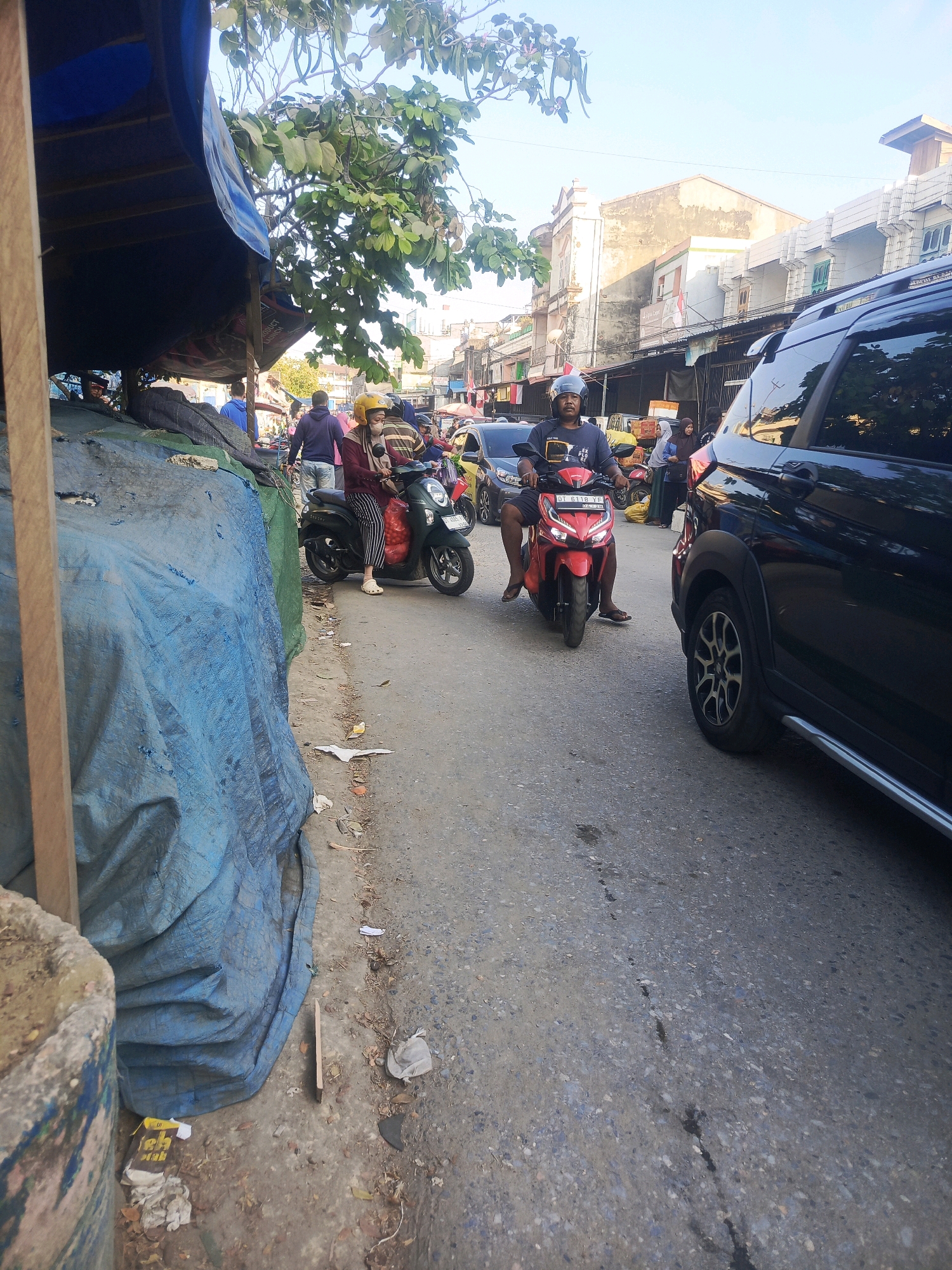 A street scene in Kendari, Indonesia, depicts a moderately busy road with several people on motorcycles and a black SUV. On the left side of the road, a blue tarp covers what appears to be market stalls or stored goods, with a wooden post in the foreground. A man wearing a helmet and a dark t-shirt is riding a red motorcycle. Further back, a woman in a yellow helmet and a patterned shirt is on a dark green scooter, with a person behind her. Buildings line the right side of the street, some appearing to be commercial establishments with balconies. The road surface is asphalt, with some debris and a stain visible. The sky is clear and blue.