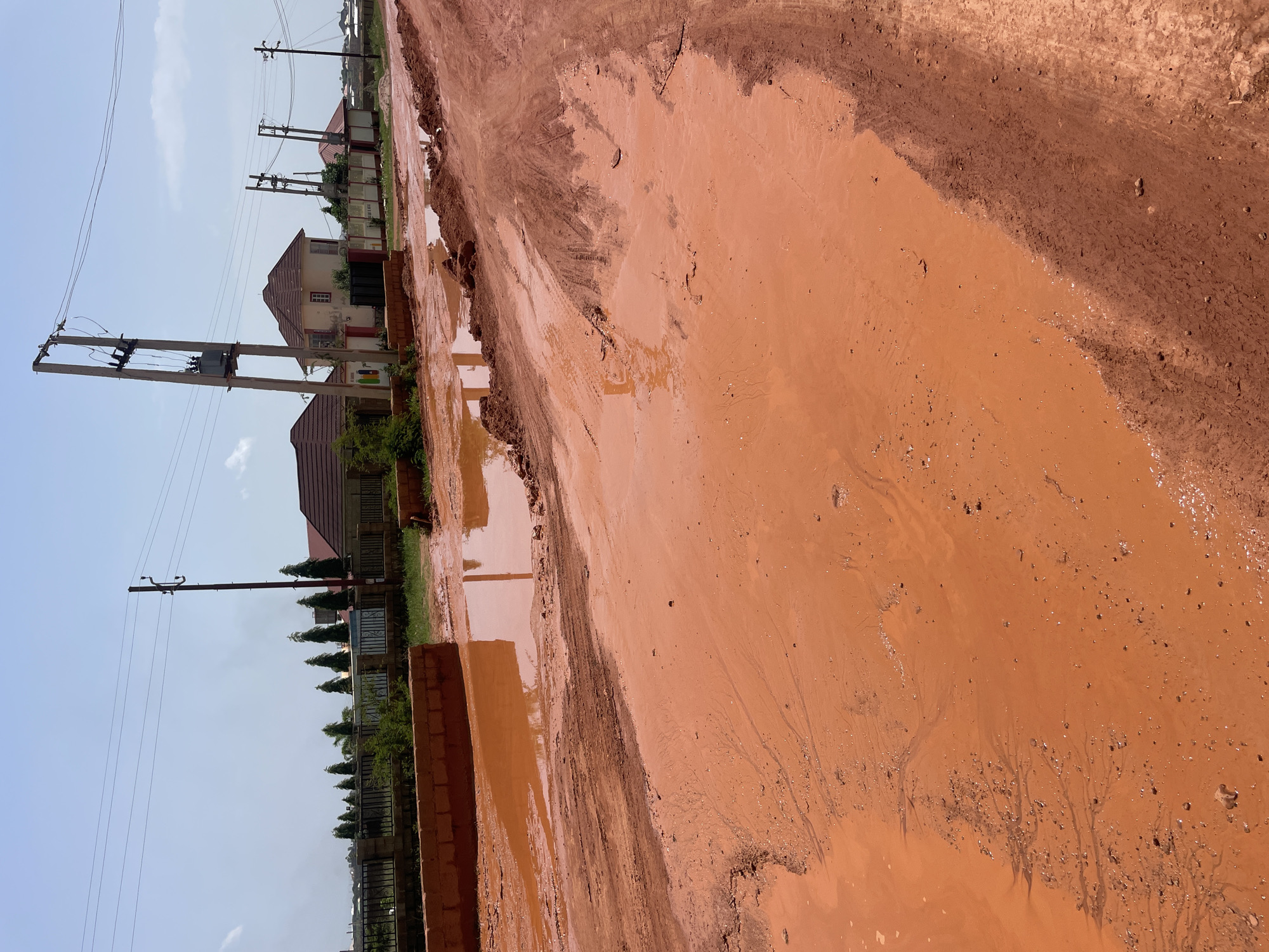 The image depicts a muddy, red dirt road in the city of Jos, Nigeria. Large puddles of muddy water have formed on the road, creating a challenging driving surface. In the background, several houses with red tile roofs are visible, along with utility poles and power lines. The sky is clear and blue, with a few scattered clouds. The overall scene suggests a rural or semi-rural setting that has been affected by recent rainfall.