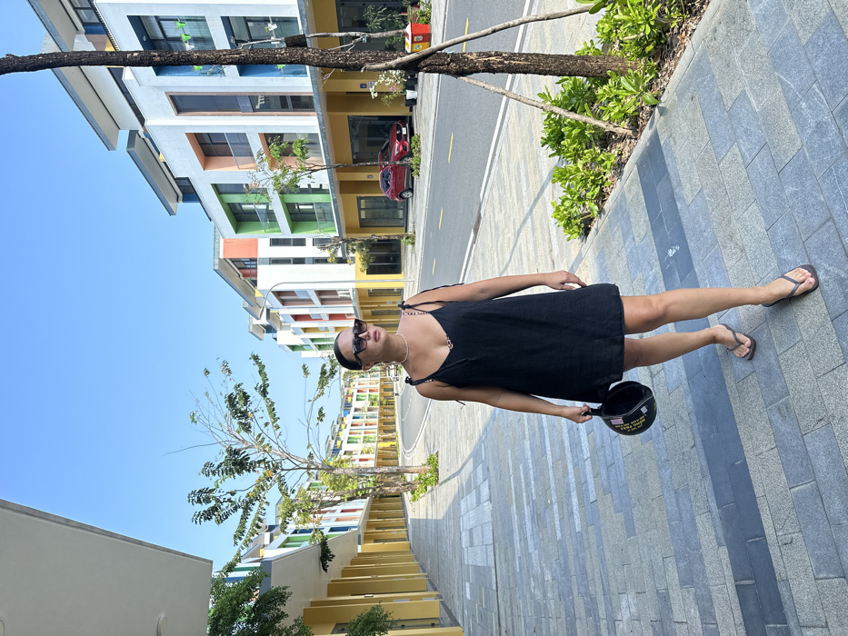 The image depicts a female subject walking on a paved sidewalk in a modern urban street in Phú Quốc, Vietnam, under a clear blue sky. The subject, an adult female with dark hair styled back, wears dark sunglasses, a white pearl necklace, a black linen-style spaghetti-strap dress with tie details on the shoulders, and dark flip-flops. She holds a black open-face helmet with white lettering in her left hand. She is in a mid-stride pose, looking towards the right of the frame.
The sidewalk is composed of light and dark grey rectangular paving slabs. To the right of the sidewalk is an asphalt road with white lane markings. Rows of multi-story, modern buildings line both sides of the street, featuring white facades with vibrant colorful accents in yellow, green, orange, red, and blue around windows and balconies. The ground floors of these buildings appear to be commercial units with large glass windows. Small trees with green foliage and shrubbery are planted along the sidewalk and adjacent to the road. A red SUV is parked on the street on the right side of the image. The scene is brightly lit by natural sunlight, casting shadows.