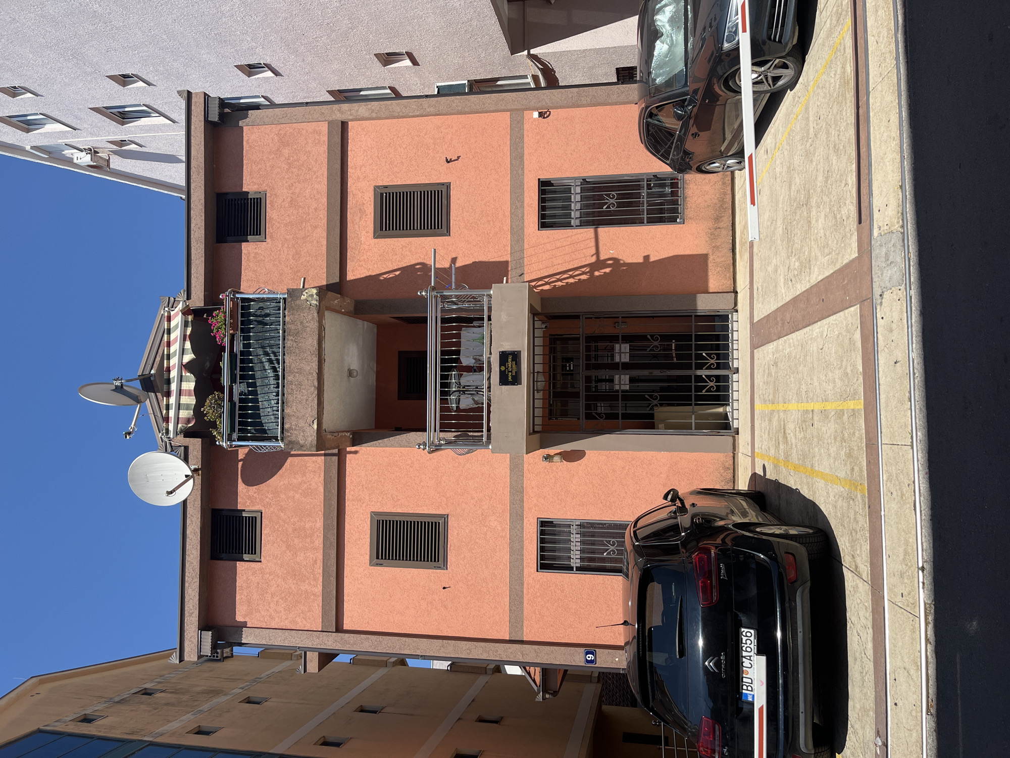 This is an outdoor, low-angle, vertical-format photograph showing the facade of a building with an orange-pink stucco finish. The building has several windows with dark metal grates. A balcony with a metal railing and some potted plants is visible on the left side, partially covered by an awning with red and white stripes. To the left of the building, a large satellite dish is mounted on the wall, and another smaller one is on the roof.
In the foreground, a black Citroen car is parked in a lot. The car's license plate reads "BD CA 656". A white barrier arm is positioned behind the car, suggesting a parking area with entry control. The ground is paved with concrete, marked with yellow lines for parking spaces. The sky is a clear, bright blue, indicating a sunny day. The architecture and the car's license plate suggest the location is Budva, Montenegro. There is no visible activity or people in the image. The overall impression is that of a residential or commercial building in a sunny, urban environment.