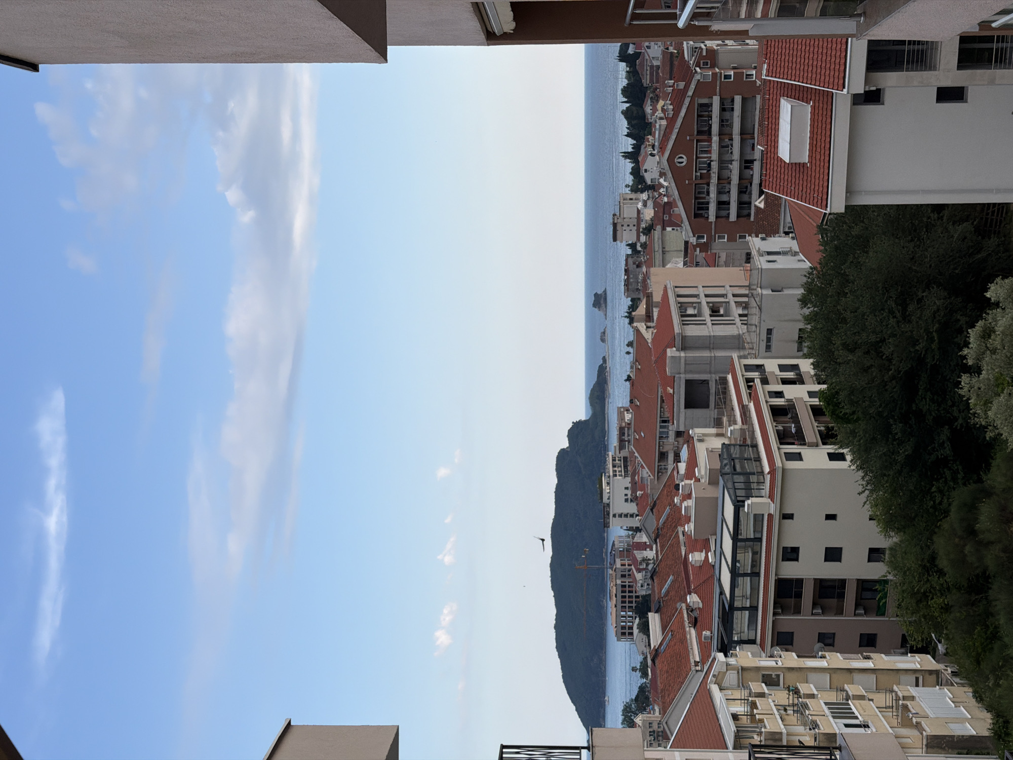 An aerial, medium shot captures a view of Budva, Montenegro, featuring a cityscape and a body of water. The sky is predominantly blue with scattered white clouds. The foreground is occupied by parts of buildings, with balconies and windows visible, framing the scene.
The midground showcases a dense arrangement of buildings, characterized by red-tiled roofs and varying architectural styles. Some buildings appear residential, with balconies and numerous windows, while others seem to be under construction or have a more commercial appearance, including one with a glass-enclosed rooftop structure. A construction crane is visible among the buildings.
The background consists of the Adriatic Sea, a large, green, mountainous island, and a few smaller rock formations emerging from the water. The sea is calm, reflecting the blue of the sky. A bird is in flight over the sea and the island. The overall impression is of a coastal city on a sunny day.