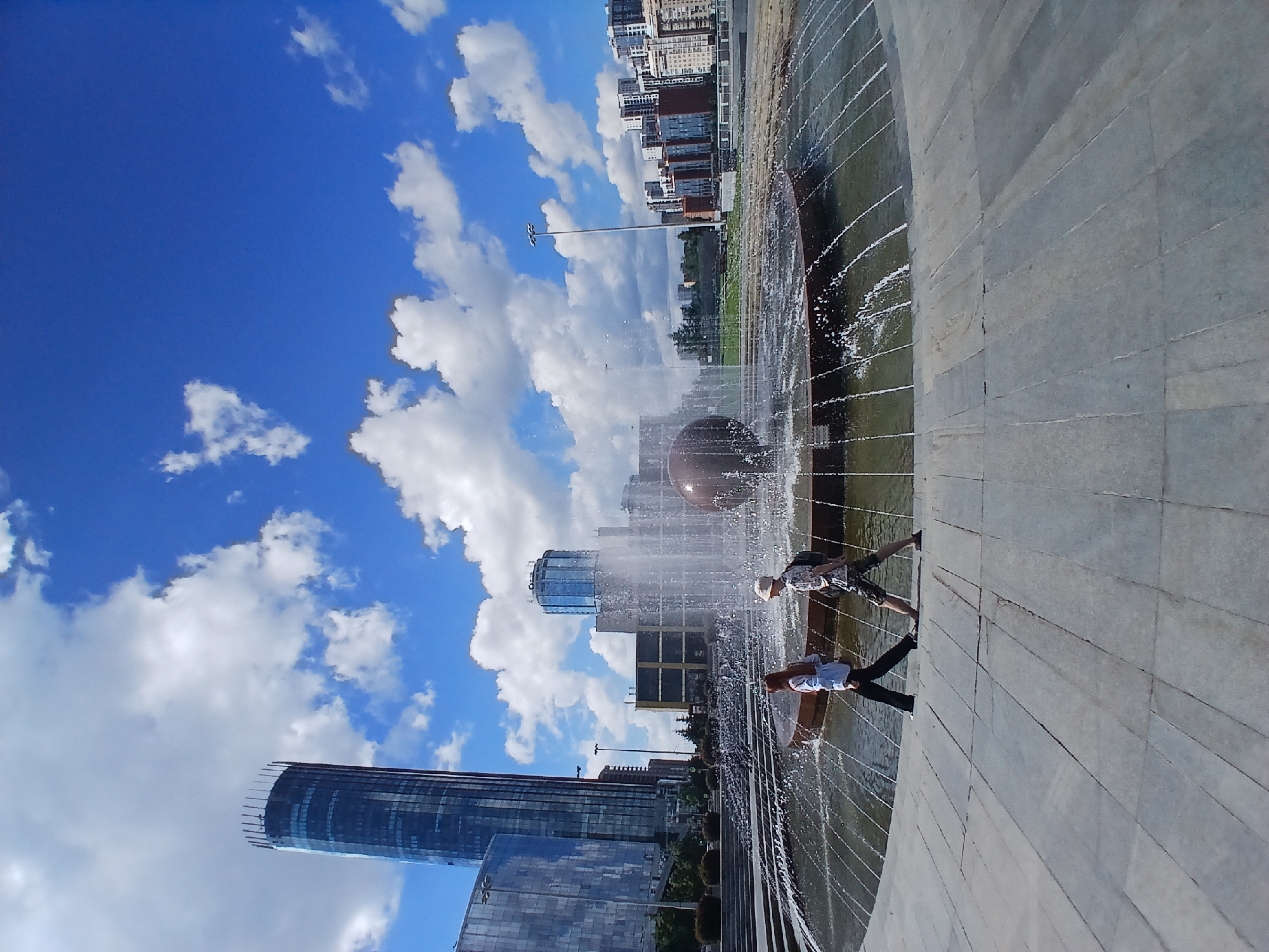 The image depicts a contemporary urban scene in Yekaterinburg, Russia. The foreground features a wide, circular fountain with water jets spraying upwards, creating a dynamic visual effect. Two individuals are visible interacting with the fountain, with one person appearing to stand on the edge and another walking on the wet surface. A large, dark sphere is submerged in the fountain's basin, partially obscured by the water.
In the background, a modern cityscape unfolds under a bright blue sky dotted with fluffy white clouds, suggesting fair weather and a daytime setting. Several tall buildings, including a distinctive curved skyscraper and other multi-story structures, dominate the skyline. The architectural styles suggest a relatively new urban development. The ground around the fountain is paved with large, grey tiles arranged in a patterned fashion. The overall impression is one of a lively public space on a clear day. There is no visible text in the image.