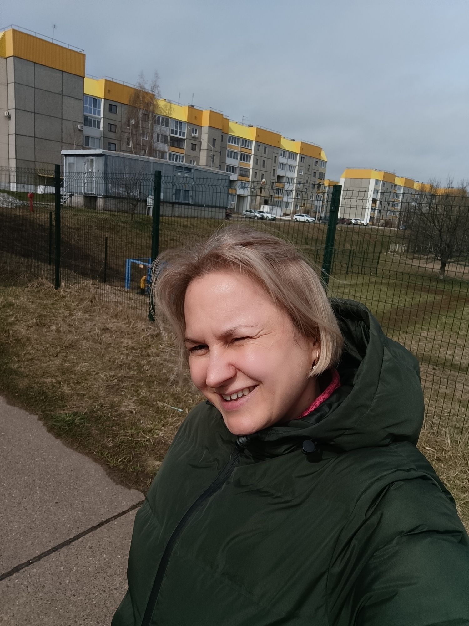 A middle-aged woman with blonde hair is smiling and winking at the camera. She is wearing a dark green puffy jacket. The background shows a row of multi-story apartment buildings with gray and yellow accents. A green metal fence separates the woman from a grassy area. A concrete sidewalk is visible in the foreground. The photo was taken in the city of Марковский, Russia.