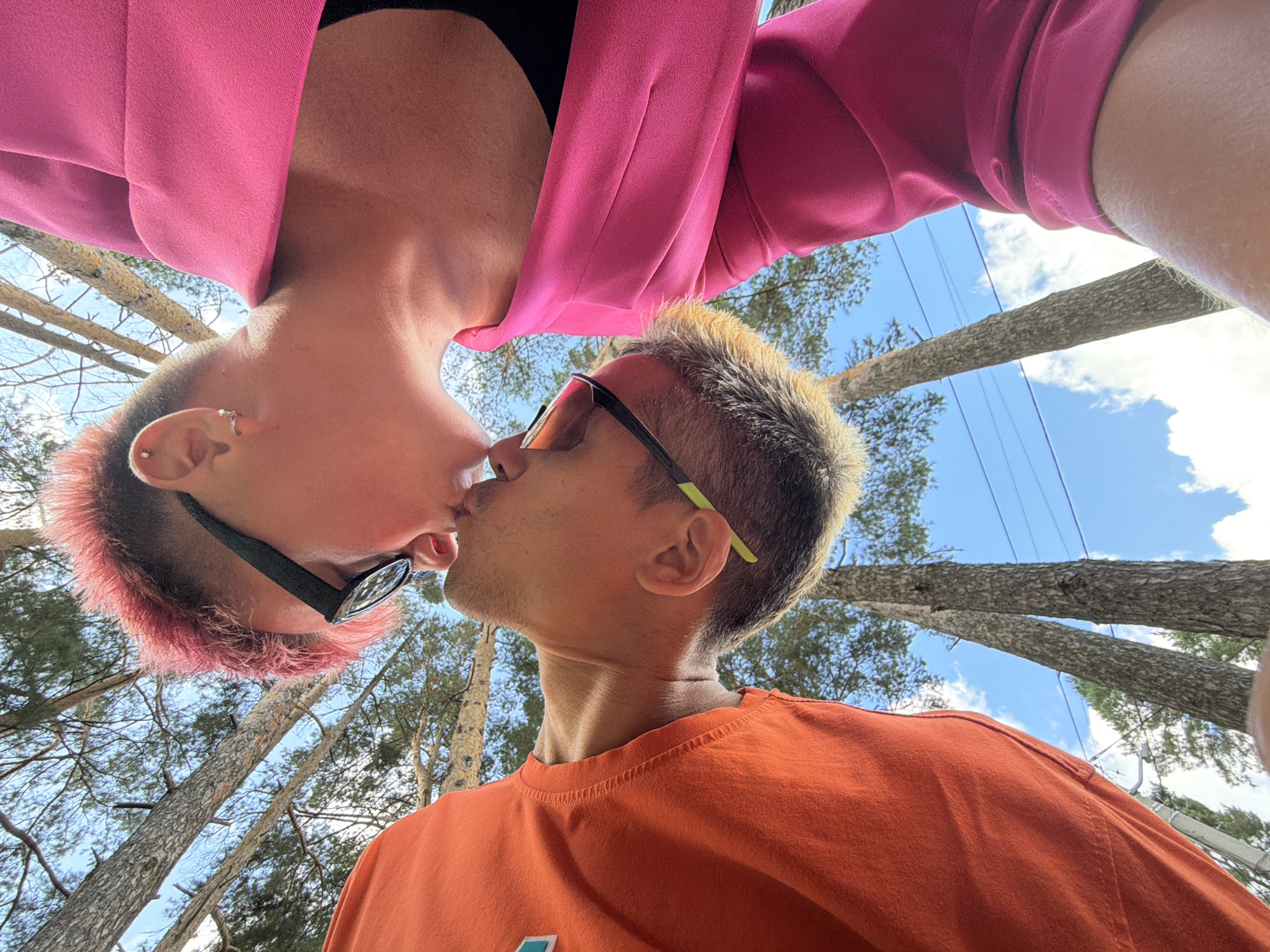 In an outdoor setting with tall trees and a blue sky with clouds, two individuals are captured in a close-up, low-angle shot. One person, on the left, wears an orange long-sleeved shirt and appears to be kissing the other person. This individual has short, spiky pink hair and is wearing black-rimmed glasses. The second person, on the right, is wearing a bright pink blazer over a black garment. Their head is tilted down, and they are also wearing glasses with yellow accents on the frame. The focus is on their faces as they lean in for a kiss, with the background of trees and sky suggesting a natural or park-like environment. The exact location within Chaikovsky, Russia, cannot be determined from the visual information provided.