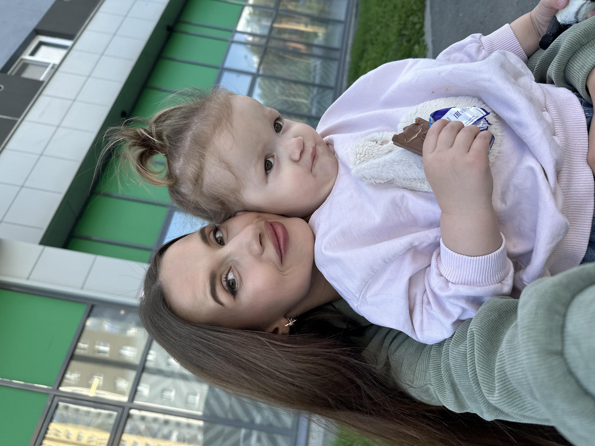 A young woman with long brown hair and subtle makeup is embracing a toddler in front of a modern building with green and gray accents. The woman is wearing a sage green hooded jacket. The toddler, dressed in a light pink sweatshirt with a fuzzy appliqué, is holding a partially eaten chocolate bar. The background features reflections in the building's windows, showing other buildings and the sky. The scene suggests an outdoor setting in an urban environment.