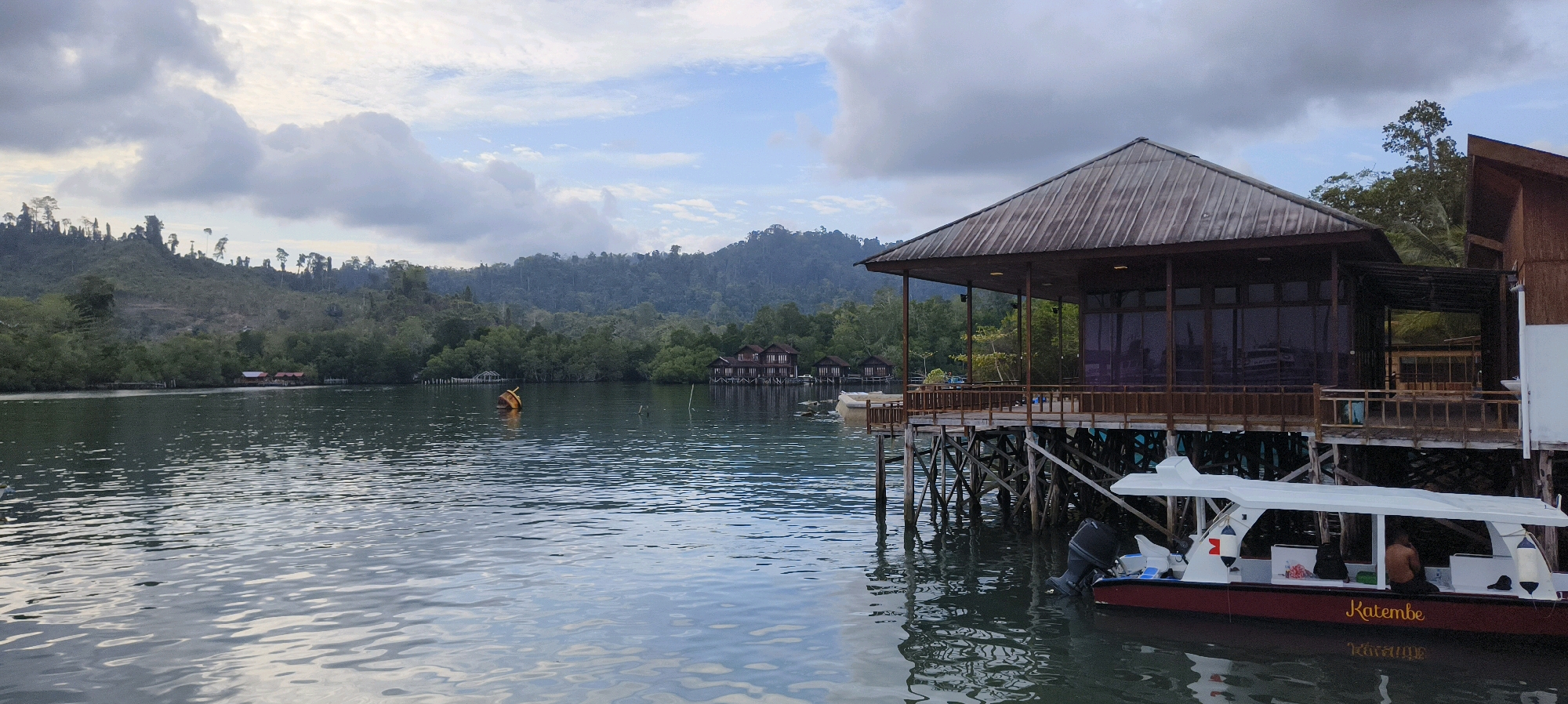A wide-angle, slightly overcast, daytime shot captures a waterfront scene with a stilted building and a boat. The foreground is dominated by calm, rippling water, reflecting the sky and surrounding structures. To the right, a wooden building with a dark, sloped roof stands on stilts over the water. It features large, dark-tinted windows and a wooden deck with railings. A white boat with a red hull is moored to the right of the building, its outboard motor visible. The name "Katembe" is visible in yellow script on the boat's hull. In the mid-ground, several similar stilted structures are visible further down the coastline, some with red roofs. A yellow buoy floats in the water between the foreground and these structures. The background consists of lush, densely forested hills and mountains under a sky with scattered clouds. The overall scene suggests a resort or accommodation in a natural, tropical environment. The location is Waisai, Indonesia.