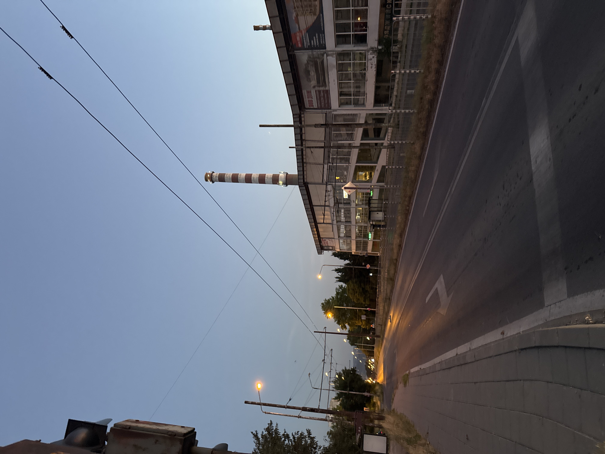 This image depicts an industrial area in Varna, Bulgaria, likely near dusk. The scene is dominated by a tall, red and white striped smokestack, a characteristic feature of industrial facilities. Adjacent to the smokestack is a long, multi-story building with numerous windows, suggesting it could be a factory or an industrial complex. The building appears somewhat dated, with advertisements visible on its facade.
The foreground is occupied by a road with clear lane markings, including a directional arrow. A sidewalk runs alongside the road, paved with grey bricks. Overhead, power lines crisscross the sky, supported by wooden poles and metal structures, indicating the presence of an electrical grid or possibly a tramway. Streetlights are visible, some illuminated, casting a warm glow onto the road, suggesting the transition from day to night. Trees and foliage are scattered along the roadside, adding a touch of nature to the otherwise industrial landscape.
The sky is a pale, clear blue, transitioning to a darker hue near the horizon, consistent with the time of day being late afternoon or early evening. There is no visible indication of specific activity or people in the image, giving it a static, observational feel. The overall atmosphere is somewhat muted, characteristic of a twilight setting in an industrial environment. No prominent text is legible in the image, although some faint lettering might be present on the building's advertisements.
