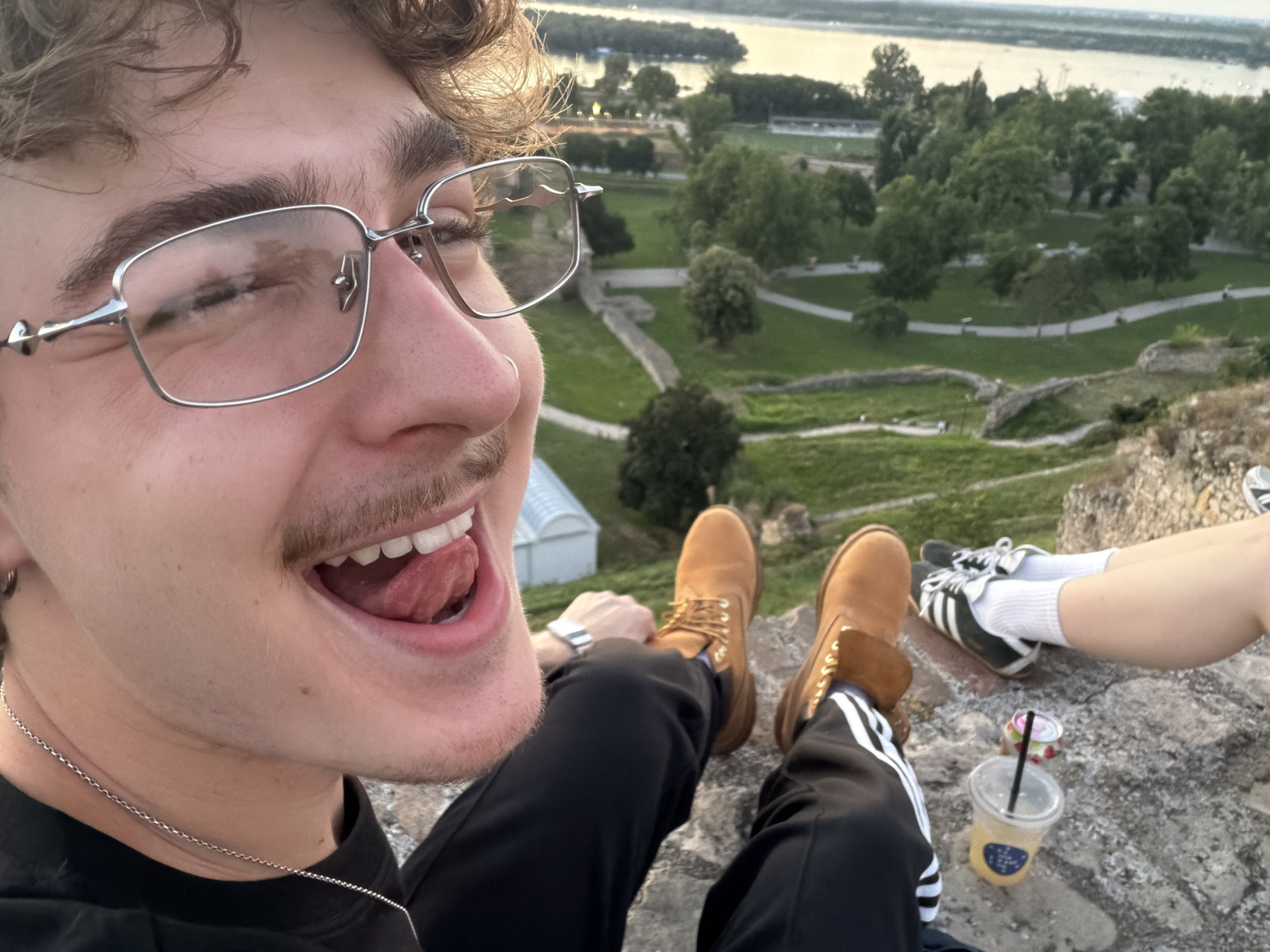 This media file is a selfie taken by a young person with curly brown hair and glasses. The subject is smiling broadly with their tongue sticking out, looking playfully at the camera. They are wearing a black t-shirt, black track pants with white stripes, and brown Timberland boots. A silver chain necklace is also visible.
In the background, the scene depicts a park with lush green grass, winding paths, and mature trees. Stone walls suggest the presence of historical ruins, possibly a fortress. A wide river is visible in the distance, reflecting the soft light of the sky. The sky appears overcast, suggesting a possibly cloudy or late afternoon/early evening time of day. Two other people's legs are visible in the lower right corner, one wearing black and white Adidas sneakers with white socks, and the other wearing similar black and white Adidas sneakers and white socks. A plastic cup with a black straw, containing a yellow beverage, and a small can are placed on the rocky ground near their feet. The overall mood is relaxed and joyful, possibly indicating a casual outing or sightseeing activity.
There is a partially visible logo on the beverage cup, but the text is not legible. The location context is Belgrade, Serbia.