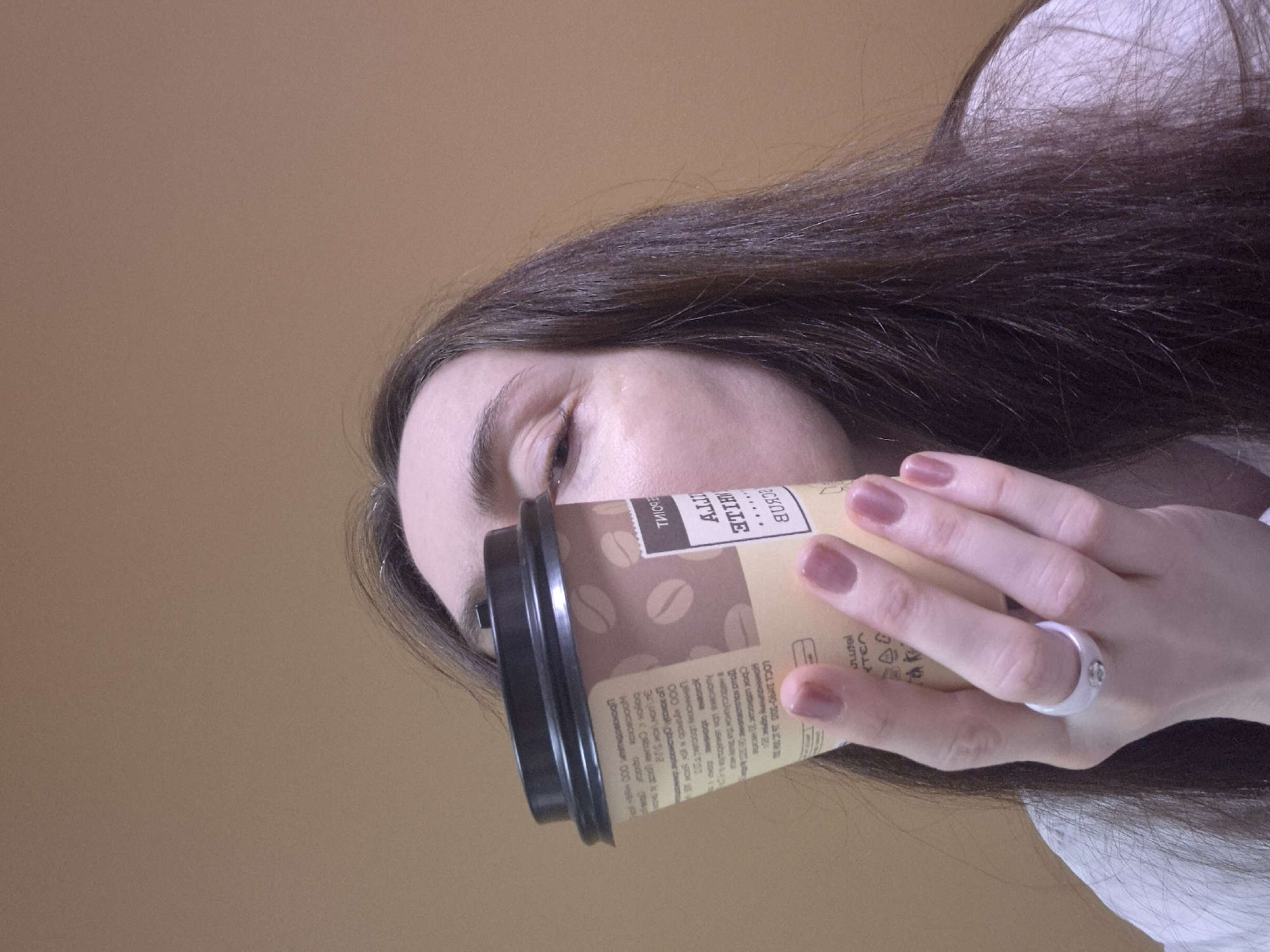 A woman is holding a coffee cup up to her face. The coffee cup is light brown with a black lid and has coffee bean graphics printed on it. Text is printed on the cup. She has long, dark brown hair and a white ring on her finger. Her nails are painted a light pink color. The background is a solid light brown color.