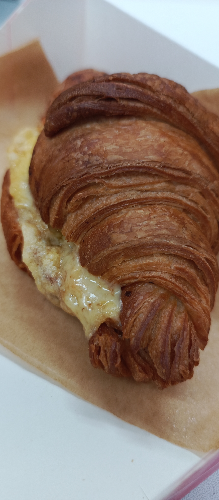 A close-up, overhead shot captures a golden-brown cheese croissant, partially opened to reveal melted, creamy cheese oozing from the center. The croissant is placed on a sheet of brown parchment paper, which rests inside a white, possibly paper, container. The background is out of focus, showing hints of white and a faint pink edge of the container. There are no people present in the image. The scene is set indoors, likely in a food establishment. The specific location in Bugry, Russia is not discernible from the visual information provided.