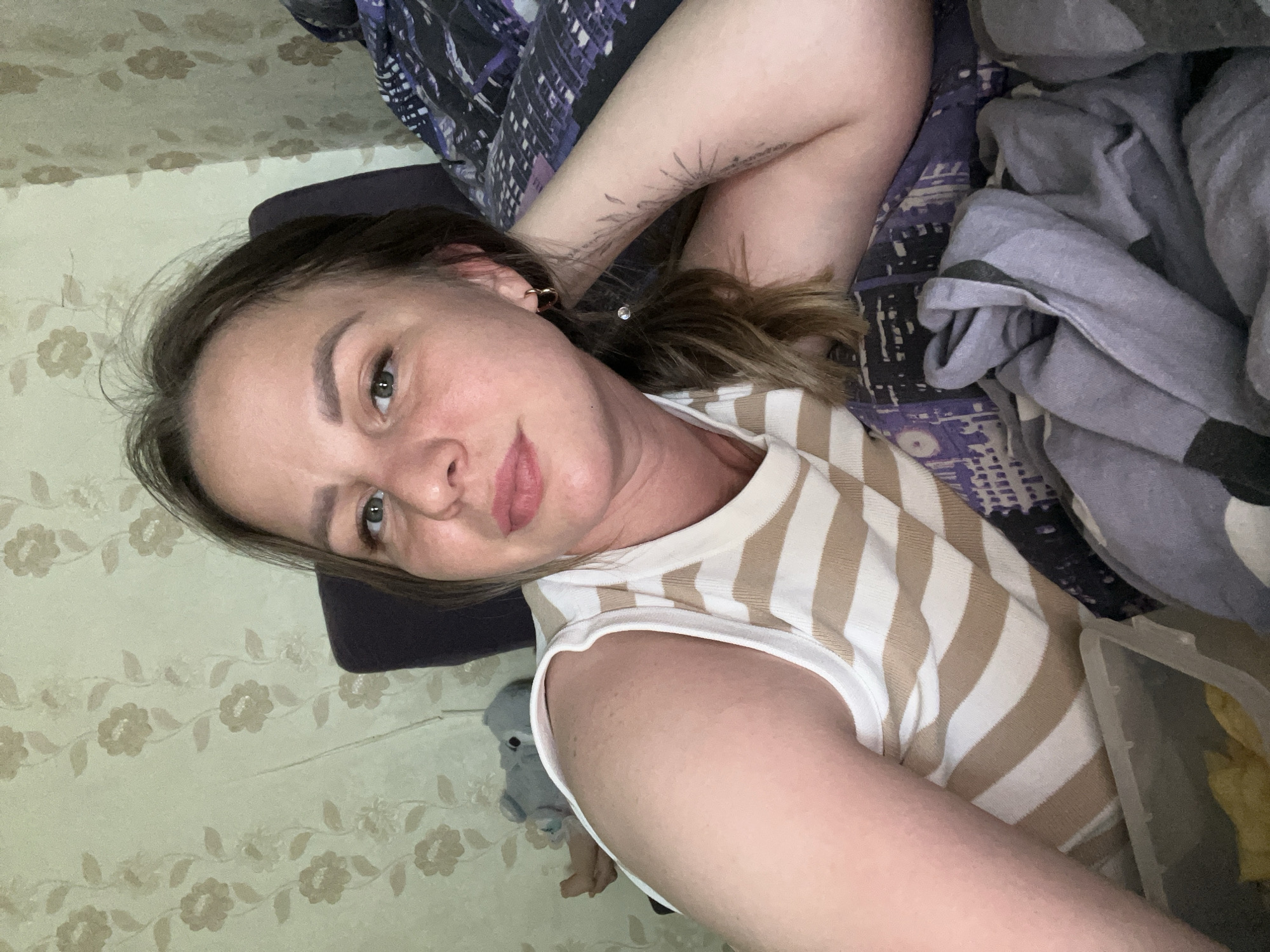 A woman lies on a bed covered with patterned bedding. She wears a striped tank top, and her hair is pulled back. Her right arm is raised above her head, revealing a tattoo. The background includes a wall with a floral pattern and miscellaneous objects. There is no indication of Izhevsk, Russia, in the image.