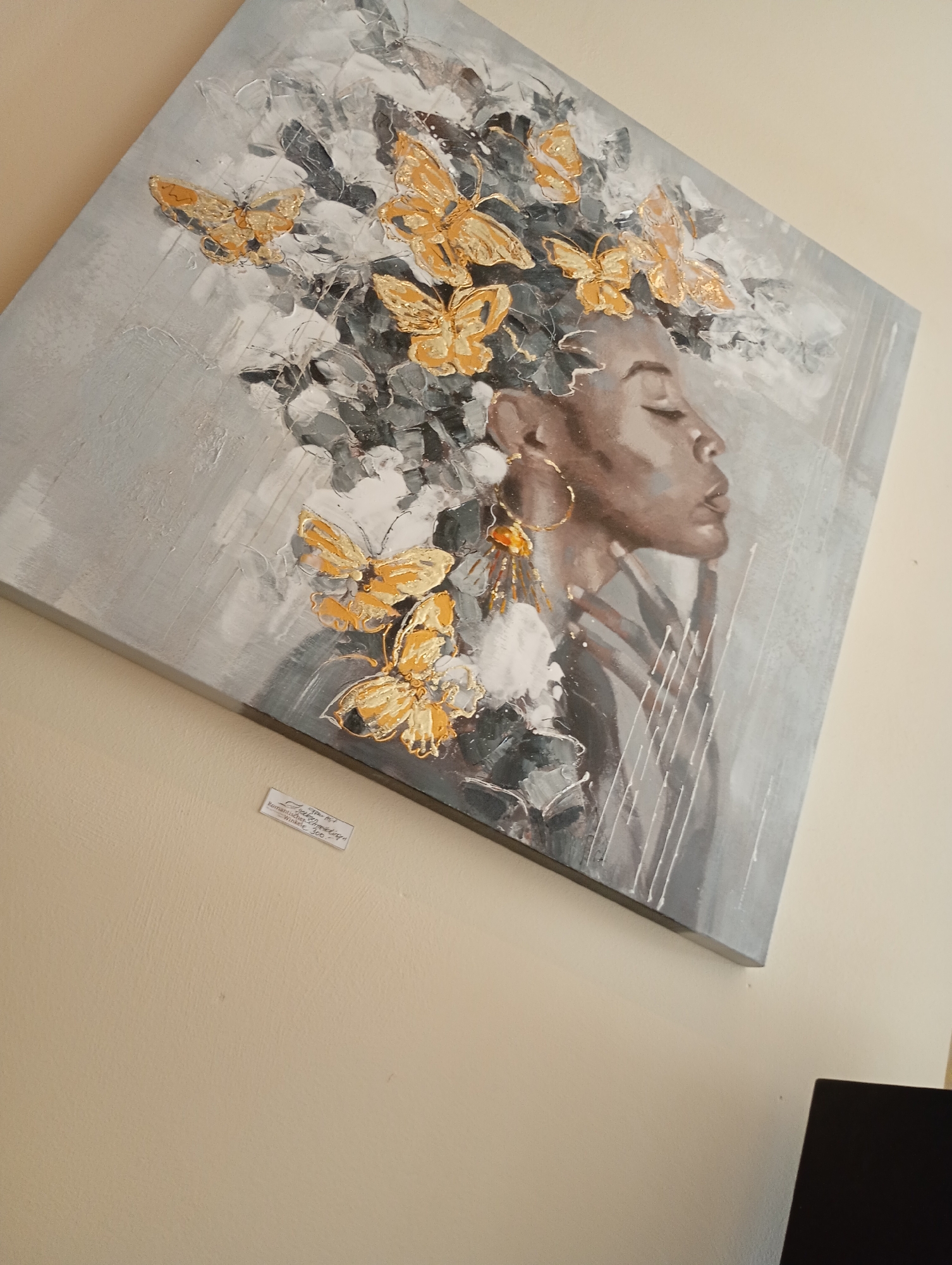 The image shows a painting of a woman with butterflies in her hair. The painting is on a canvas and is displayed on a wall. There is a small label attached to the wall below the painting. The label appears to be in German and may contain information about the painting or its artist. The text on the label is difficult to read, but some words that are visible include "Glam Art," "Romantische Schmetterlings," and "Winkels 300." It is possible that this painting is part of an exhibition or display in the town of Bad Sachsa, Germany.