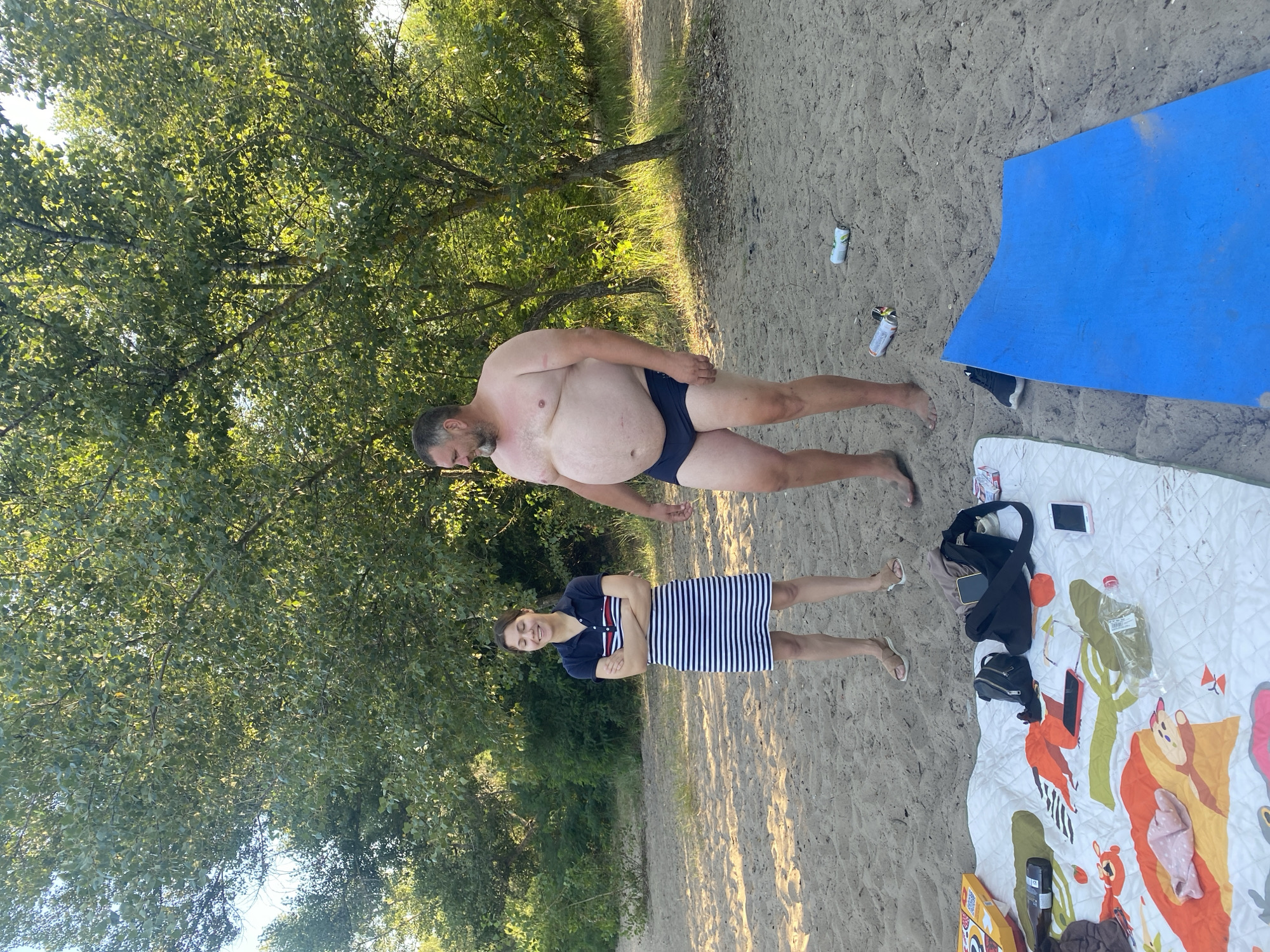 A man and a woman stand on a sandy beach under a tree in Dnipro, Ukraine. The woman is wearing a horizontally striped dress and has her arms crossed. The man is wearing black swim trunks and is looking to his left. In the foreground, a picnic blanket is spread out on the sand with various items, including a bag, phones, and a bottle of water. Several cans are scattered on the sand near the blue object in the lower right corner. The background consists of trees and foliage.