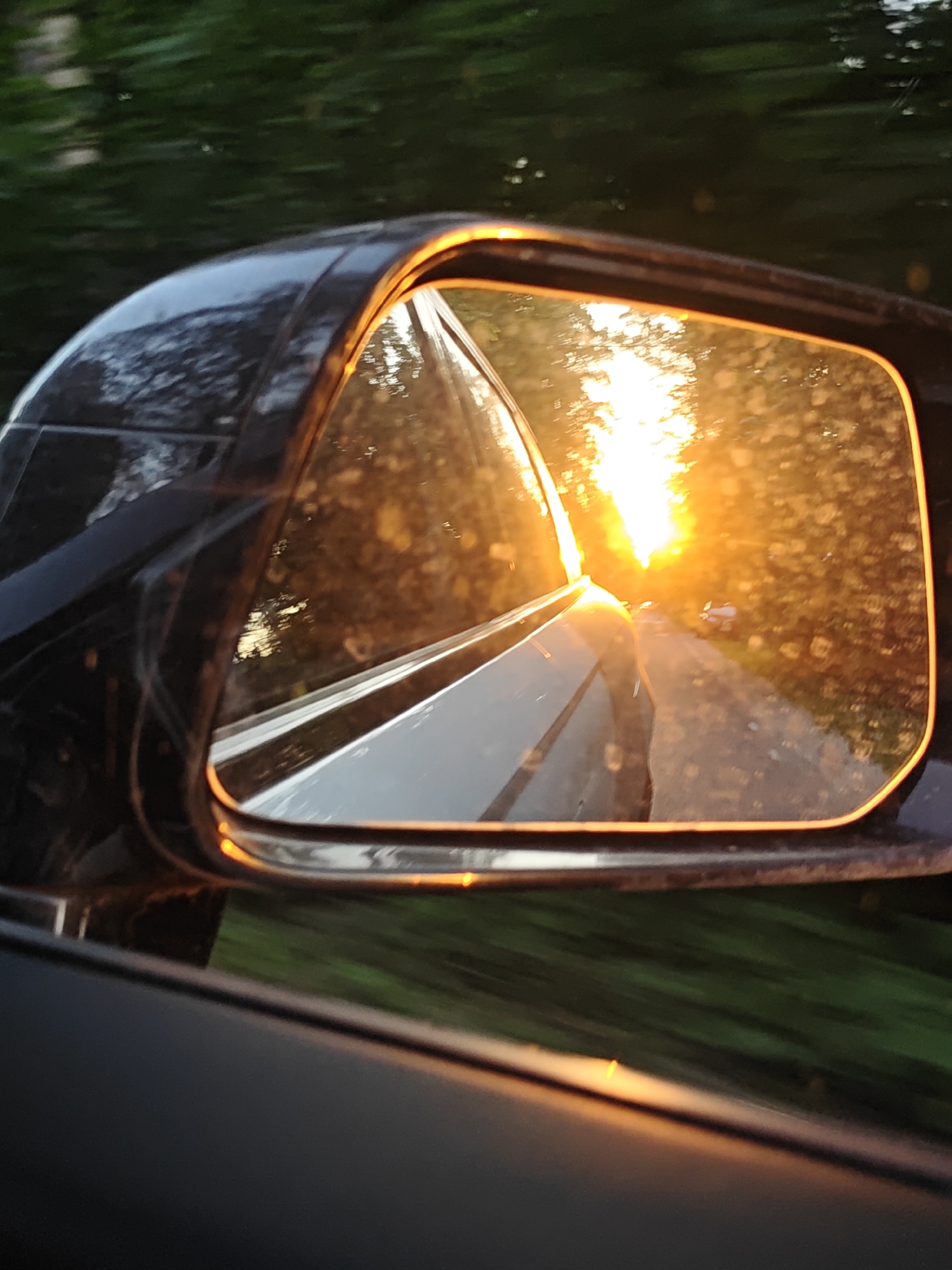 The image depicts a close-up of the passenger-side rear-view mirror of a dark-colored vehicle, likely black, in motion. The mirror reflects an intense, low-angle sun, characteristic of either a sunrise or sunset, centered on a straight, two-lane road receding into the distance.
Within the mirror's reflection, the sun appears as a brilliant, almost blinding, orange-yellow light source. The road surface is dark, suggesting asphalt or pavement, and is flanked by dense foliage and trees that are heavily backlit and tinted in warm orange and yellow hues by the sun. The mirror surface itself shows numerous small, bright speckles, indicative of dust, dirt, or water droplets illuminated by the direct sunlight. A faint reflection of the vehicle's own body, appearing metallic silver or light grey, is visible on the left side of the mirror. A small, light-colored object, possibly a mailbox or roadside marker, is discernible on the right side of the reflected road.
Outside the mirror, the glossy, dark exterior of the car's body and a portion of its window frame are visible in the foreground. The background outside the mirror consists of significantly blurred green foliage, strongly indicating that the vehicle is in motion at the time the photograph was taken. The overall environment is rural or semi-rural, characterized by a road and extensive tree cover. The image provides no visual indicators such as landmarks, signage, or specific architecture to confirm the requested location in the city of Kratovo, Russia. No people are visible in the scene.