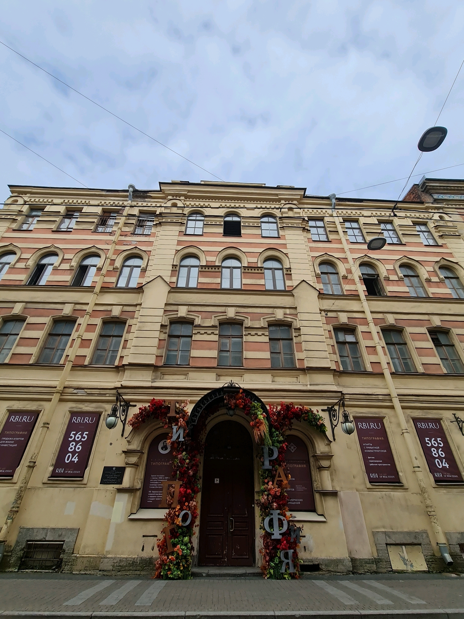 The image depicts the facade of a historic building in Saint Petersburg, Russia, characterized by its light yellow and coral striped exterior. The building features multiple stories with evenly spaced windows, many of which are arched on the upper floors. A prominent vertical pipe runs down the side of the building.
At street level, the entrance is framed by an elaborate floral archway adorned with red and orange flowers. Large, metallic letters spelling out "ПИОНЕР" (Pioner) are incorporated into the floral decoration. Several posters are visible on the building's facade. Two posters on the left side are dark purple with white text, one displaying "RBI.RU" and the other with the number sequence "565 86 04". Another poster on the right side, also dark purple, has "RBI.RU" and text related to typography and real estate.
A pedestrian crossing is visible on the paved street in front of the building. The sky above is overcast with visible clouds and thin electrical wires crossing the frame. A street lamp is also visible, suspended from wires overhead. The architectural style suggests the building is located in a historical district of Saint Petersburg.