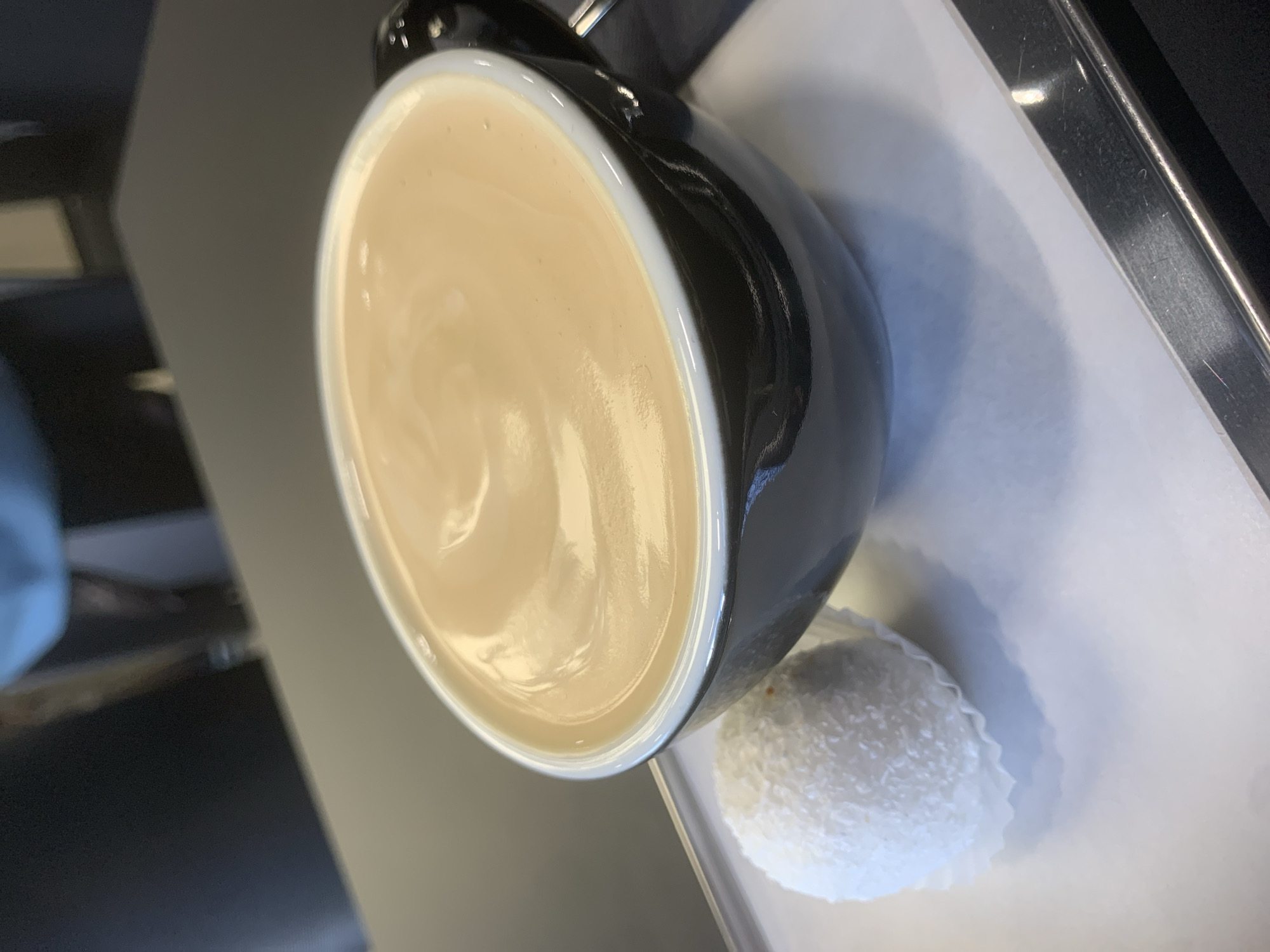 A black ceramic cup filled with a beige liquid (likely coffee with milk or cream) rests on a white surface next to a round, white, shredded coconut-covered candy in a small paper cup. The scene is indoors. The background is blurred and not clearly identifiable, so providing the specific location in Izhevsk, Russia, is impossible.